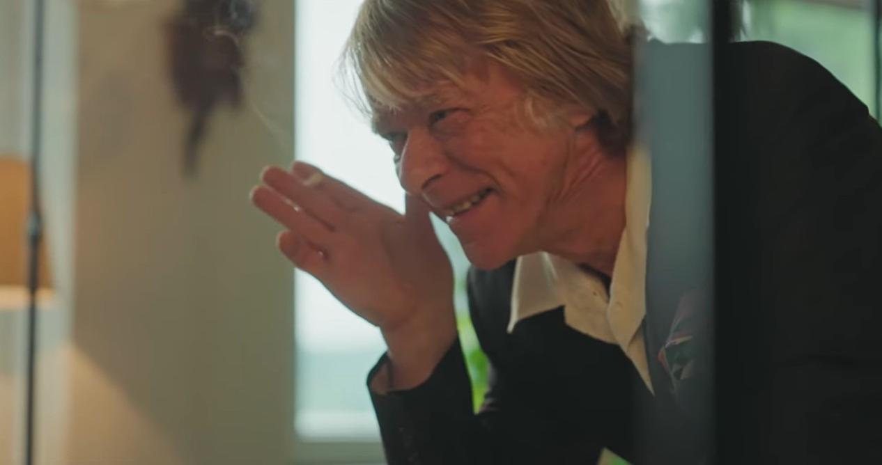 Mann mit blonden Haaren in schwarzem Anzug lächelt und hält die Hand vor sein Gesicht.