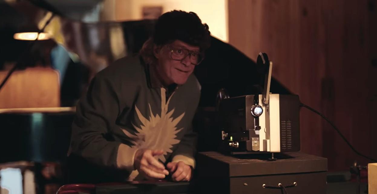 Ein Mann mit lockigem Haar und Brille vor einem Projektor.