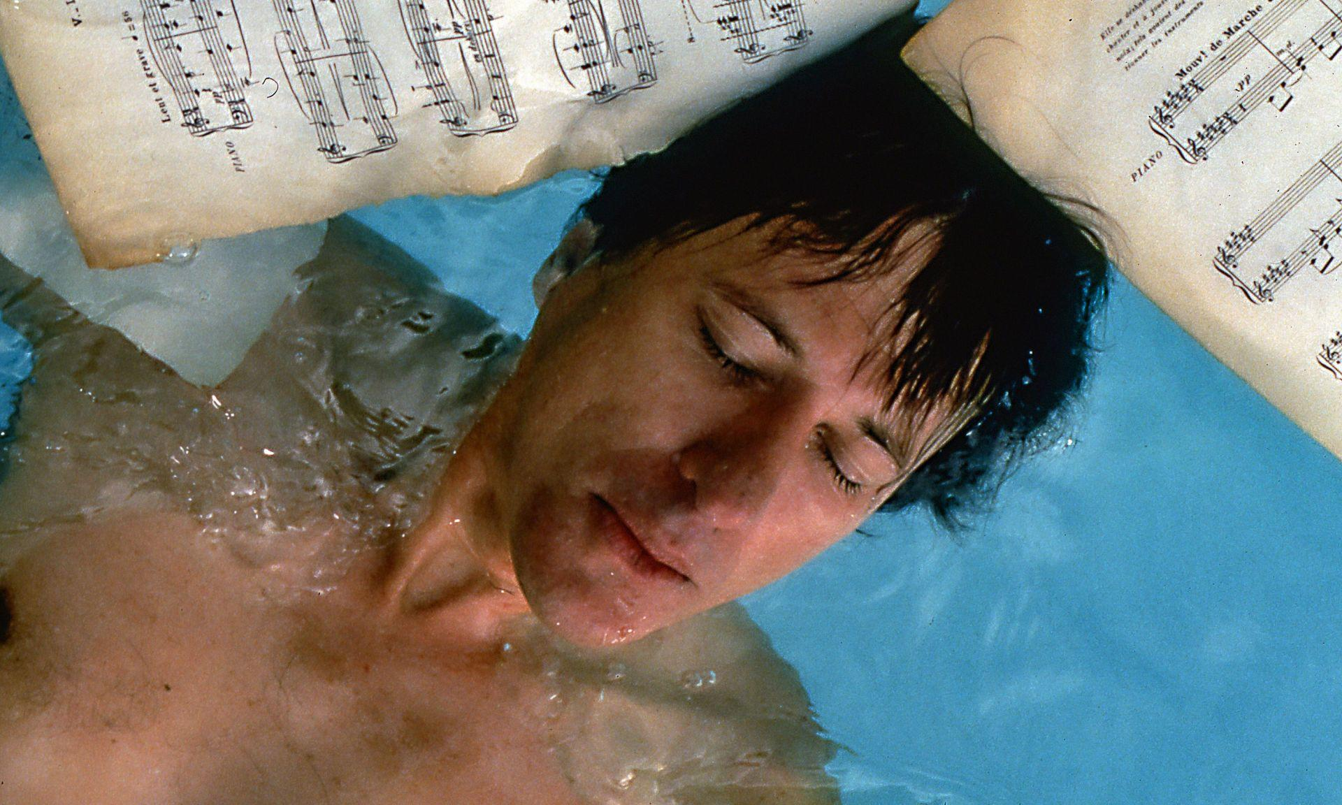 Mann in Wasser mit Notenblättern. Geschlossene Augen, braune Haare.