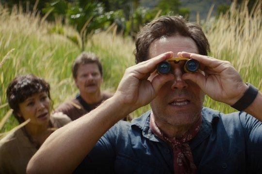 Ein Mann blickt durch Fernglas, zwei Personen im Hintergrund im hohen Gras.