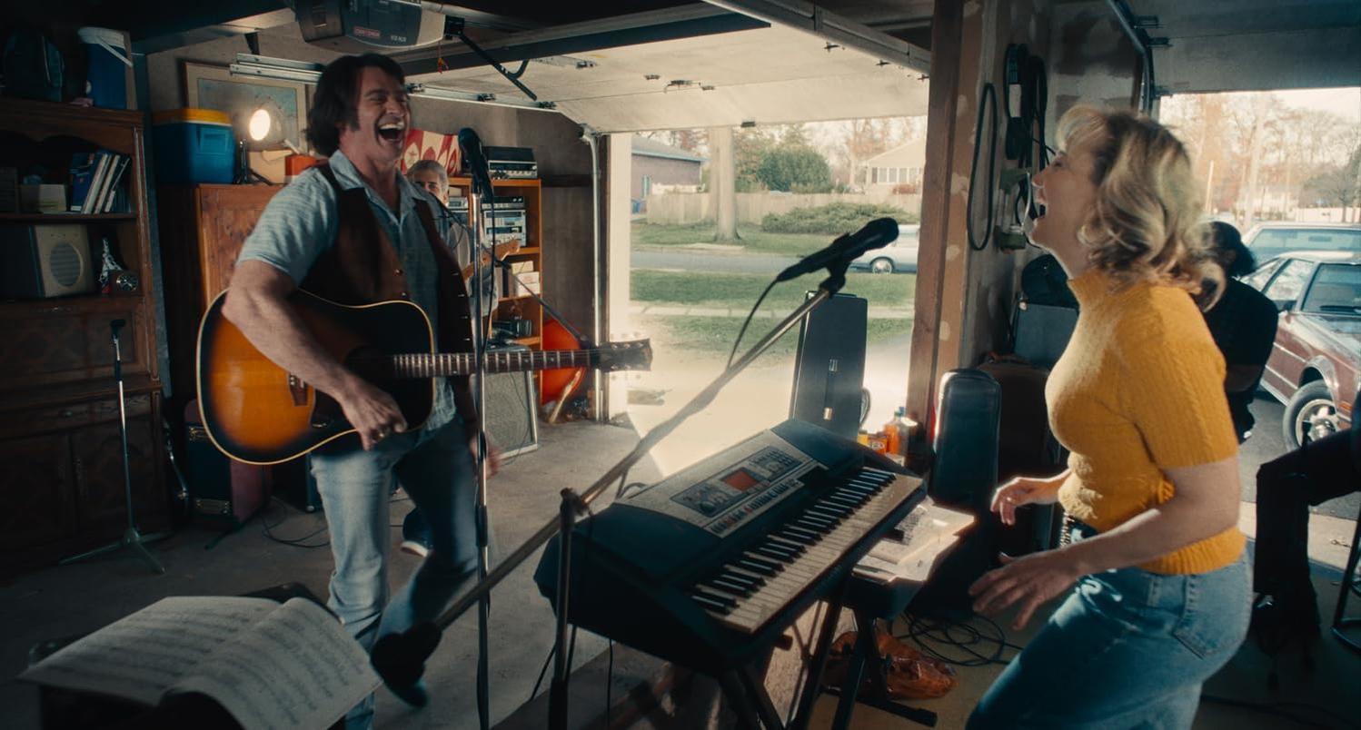 Ein Mann spielt Gitarre, eine Frau singt. Sie sind in einer Garage mit einem Keyboard und einem Garagentor.