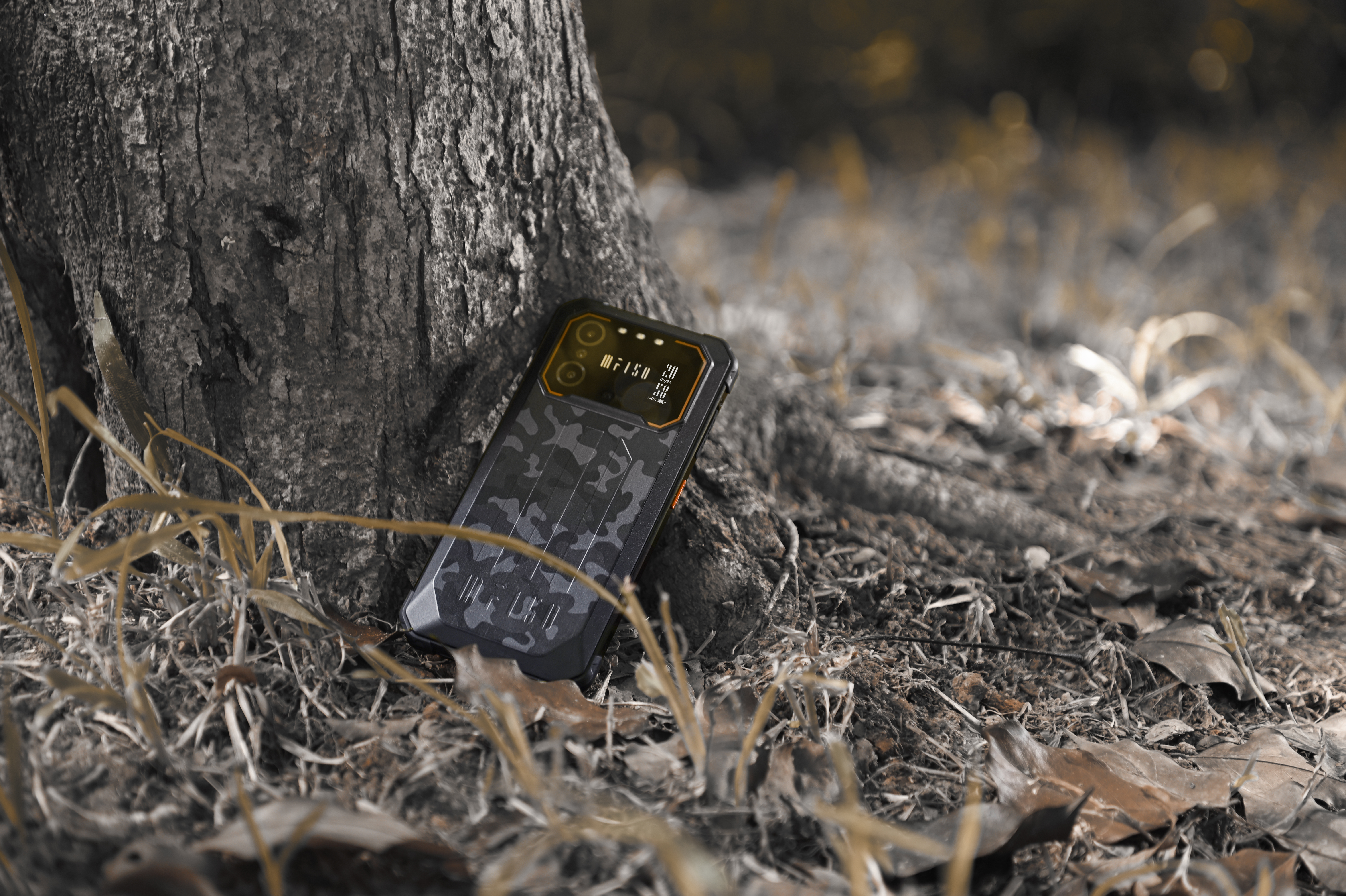 Wytrzymały telefon w kamuflażu leży obok pnia drzewa na ziemi.