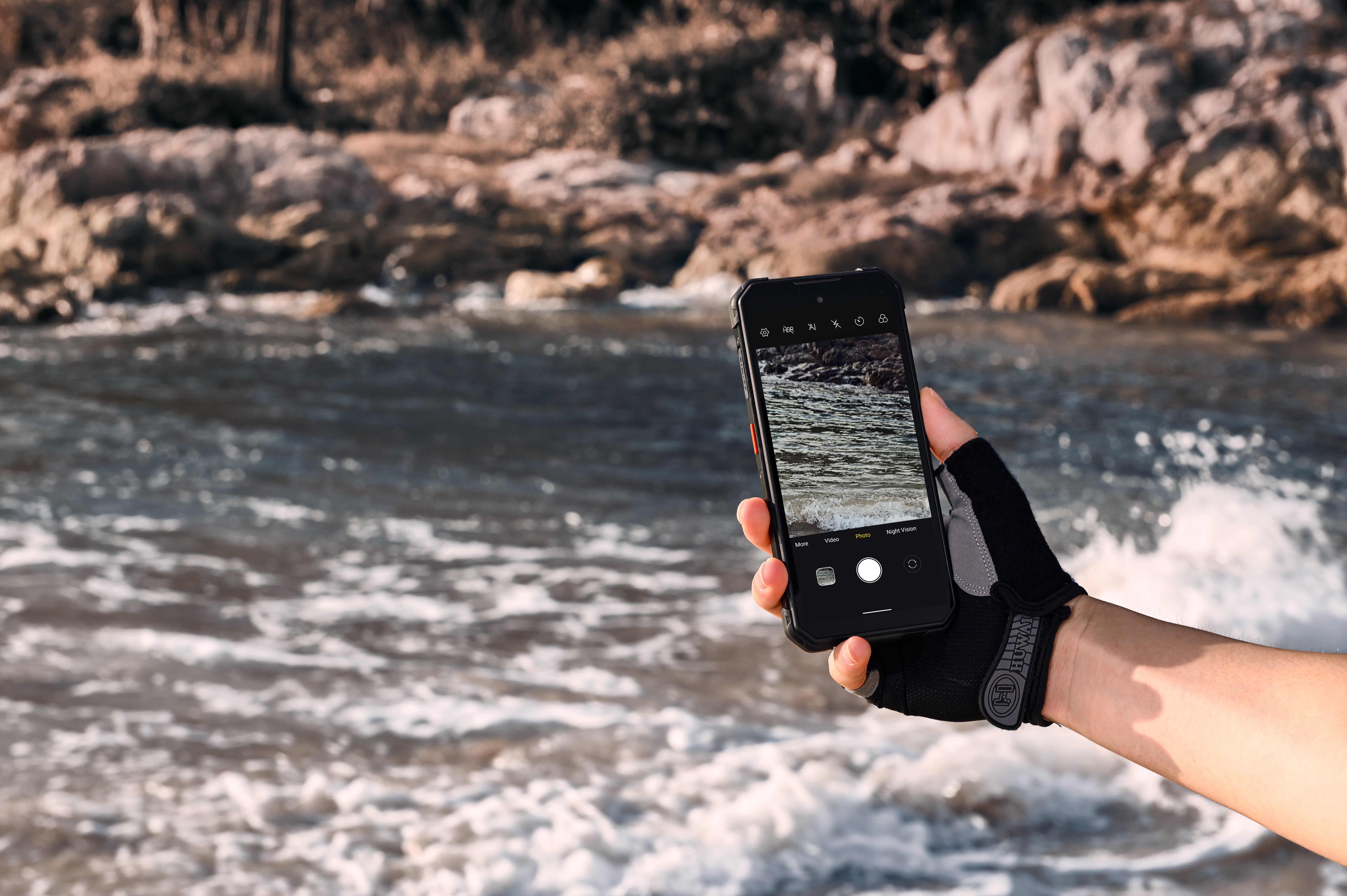 Dłoń w rękawiczce trzyma smartfon przed wzburzoną rzeką ze skałami.