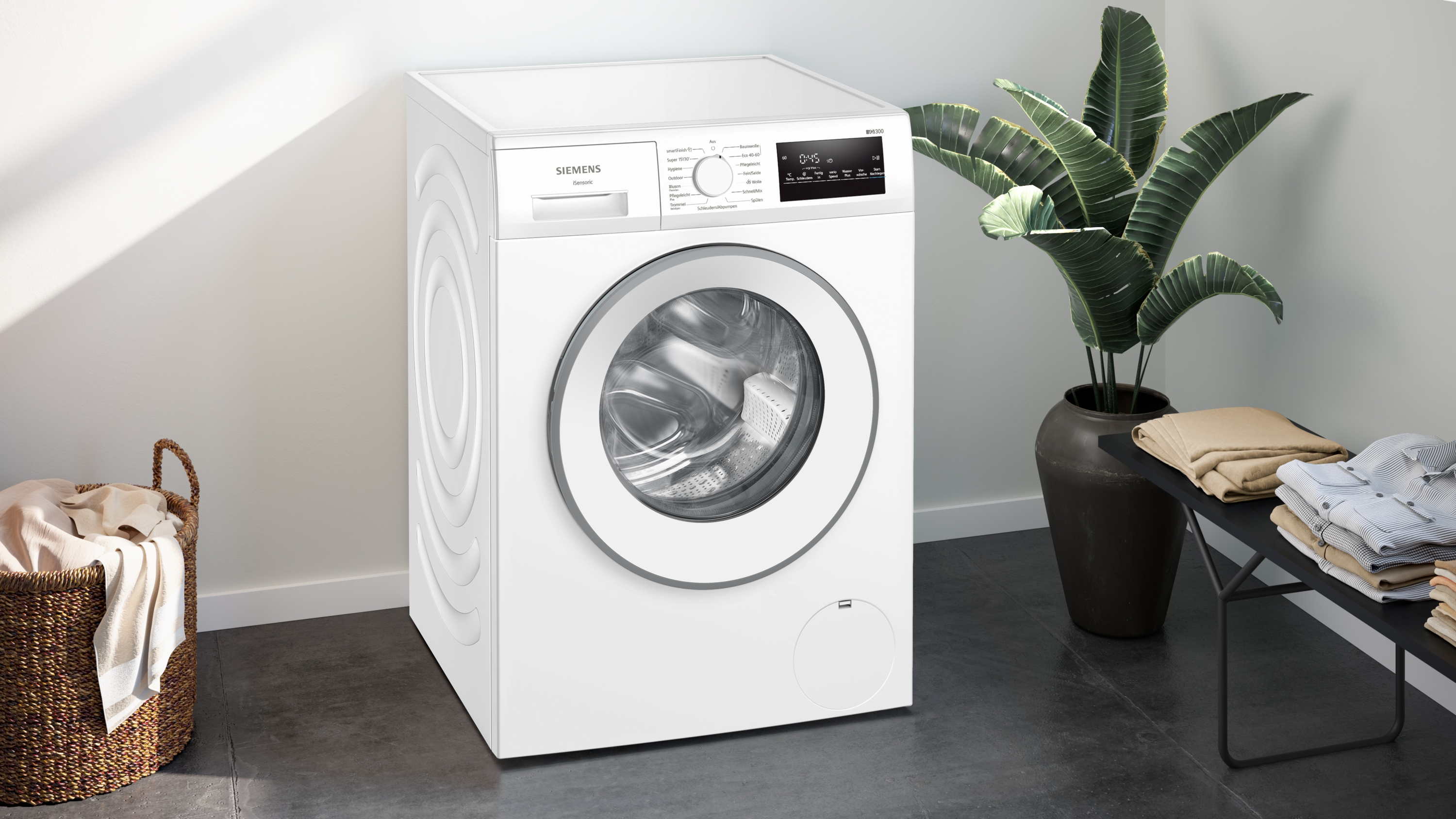 Eine weiße Siemens Waschmaschine steht neben einem Korb mit Wäsche und einer Pflanze.