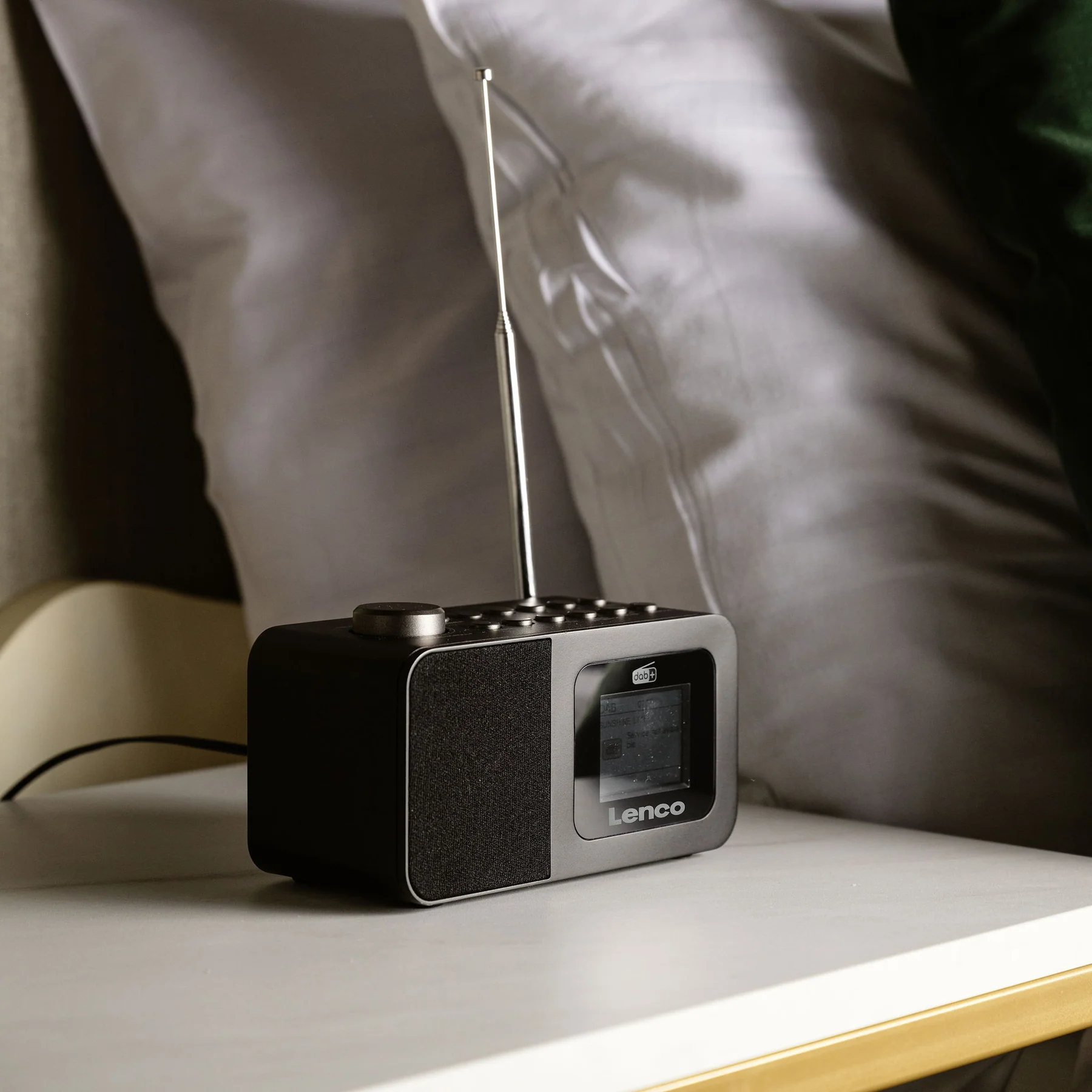 Une radio DAB+ noire avec une antenne est posée sur une table de chevet à côté de coussins blancs.