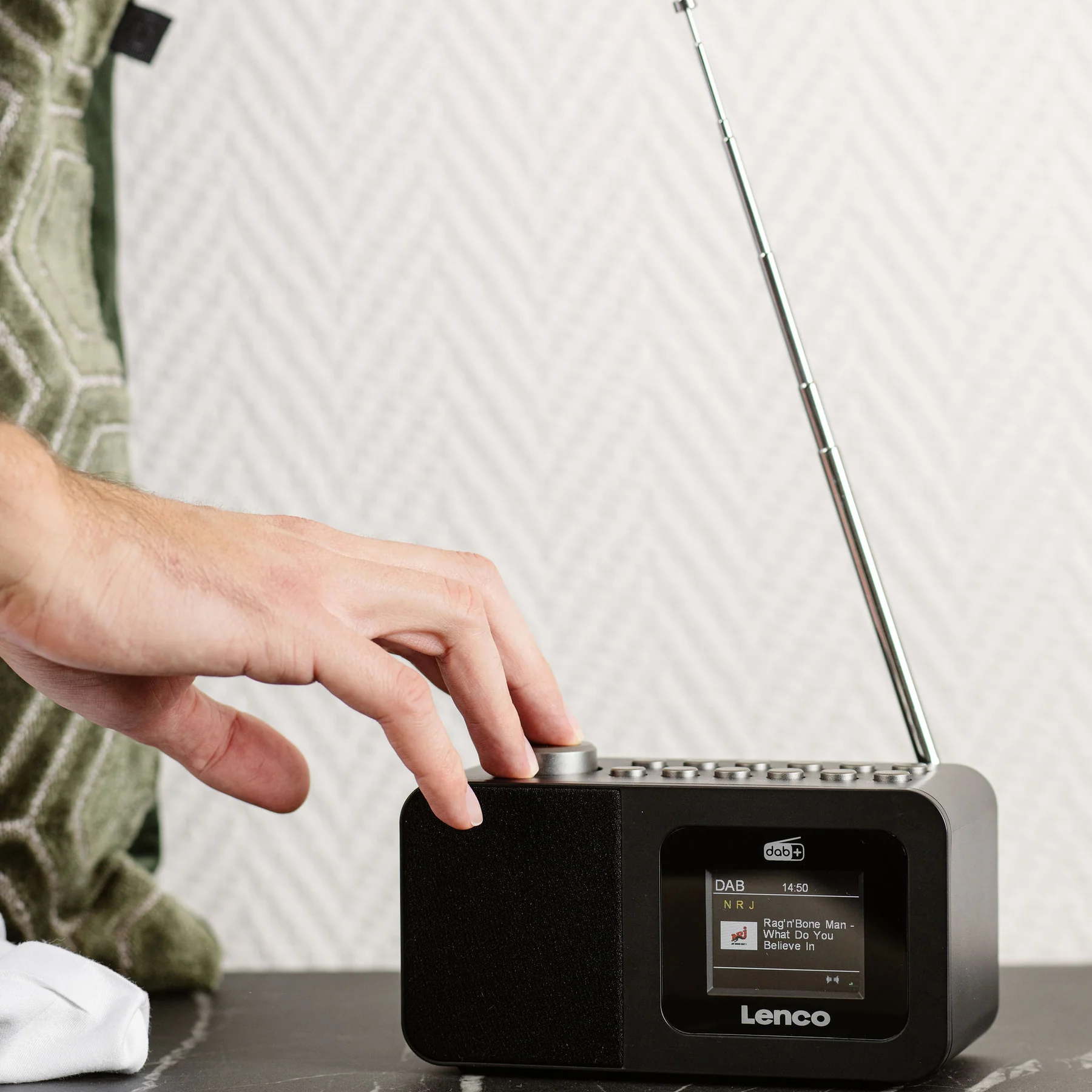 Une main tourne le bouton d'une radio Lenco DAB+ noire avec son antenne déployée.