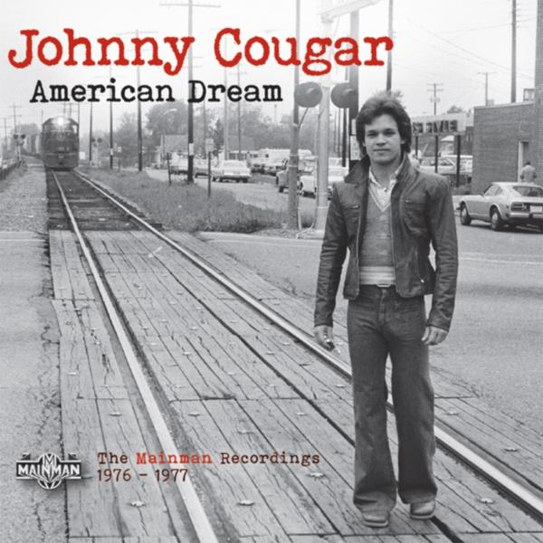 Johnny Cougar steht auf Bahngleisen. Zug und Autos im Hintergrund.