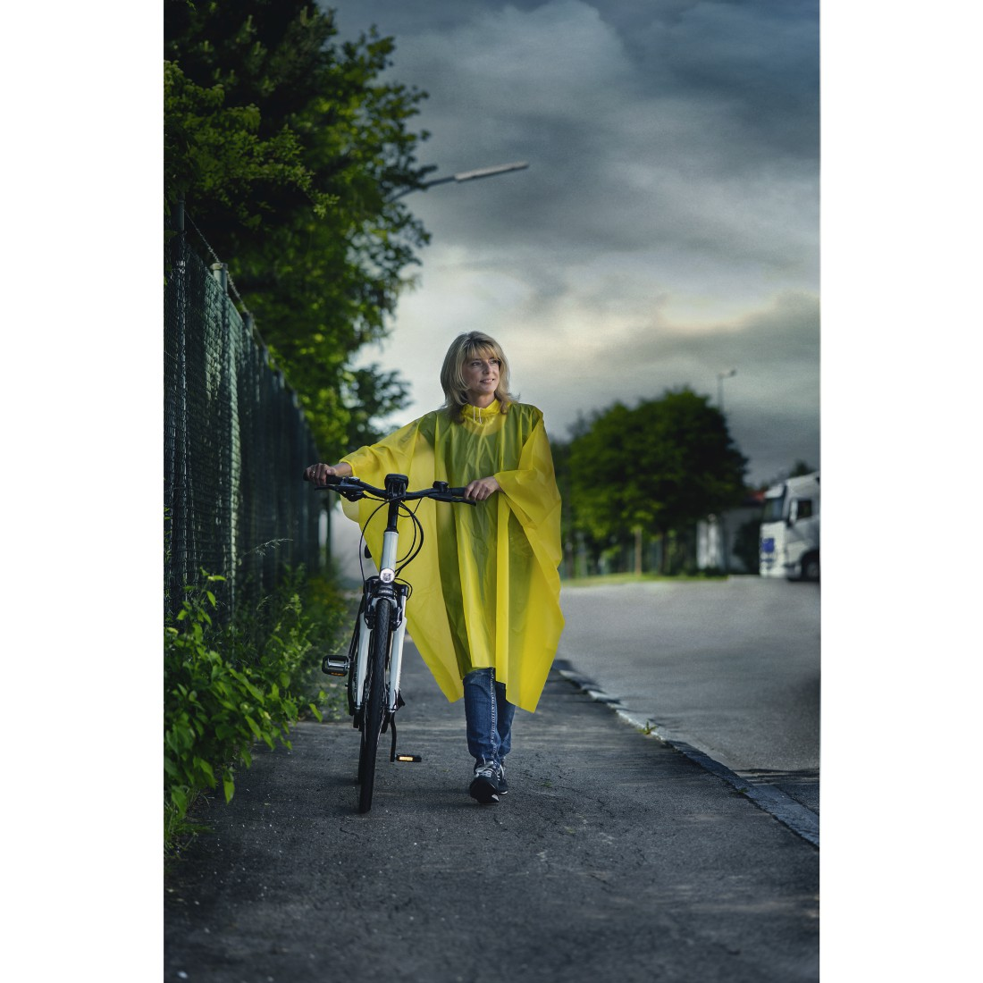 Eine Frau im gelben Poncho schiebt ihr Fahrrad auf einem Gehweg.
