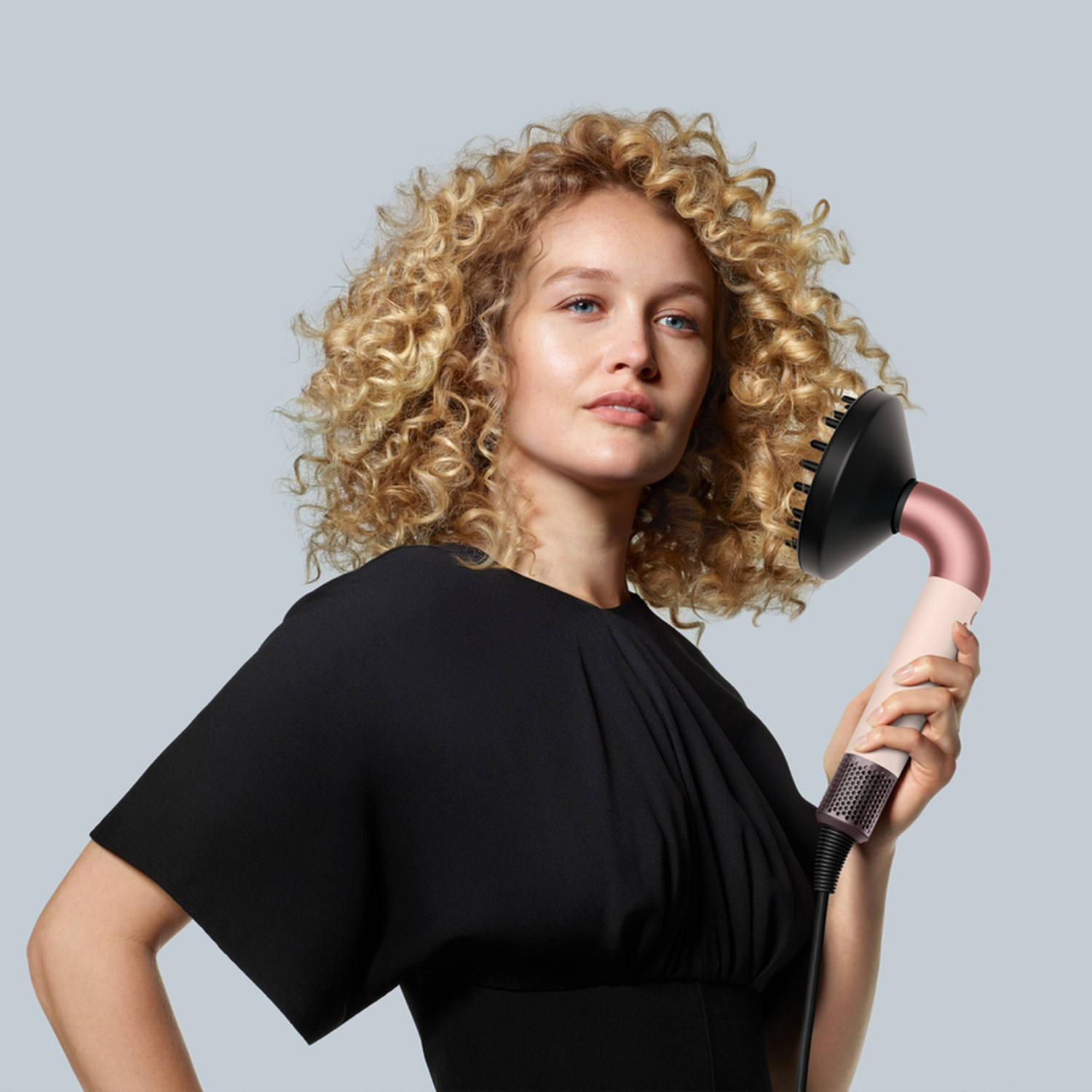 Femme aux cheveux blonds bouclés tient un sèche-cheveux rose avec diffuseur noir.