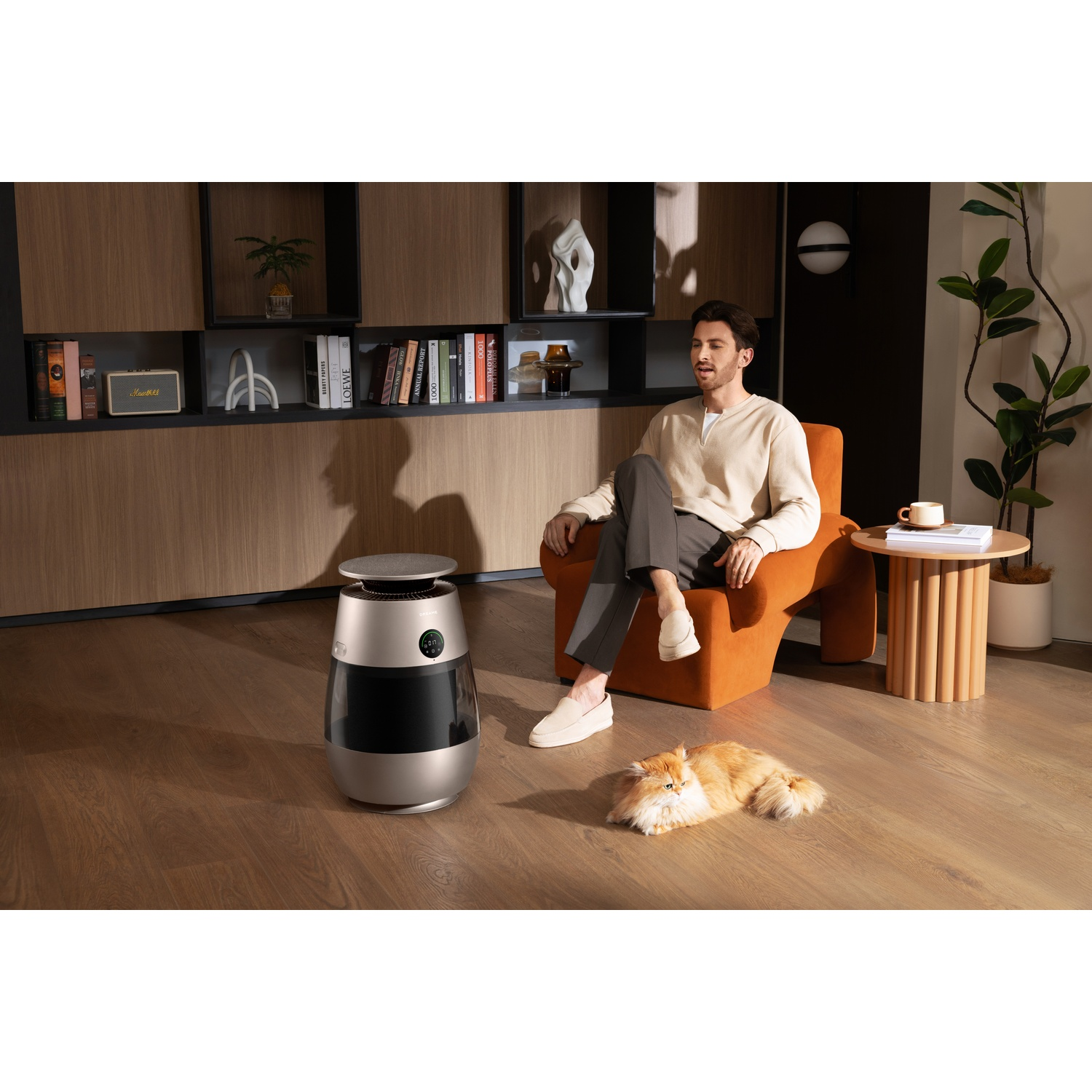 A man sits in an orange chair next to an air purifier and a cat.
