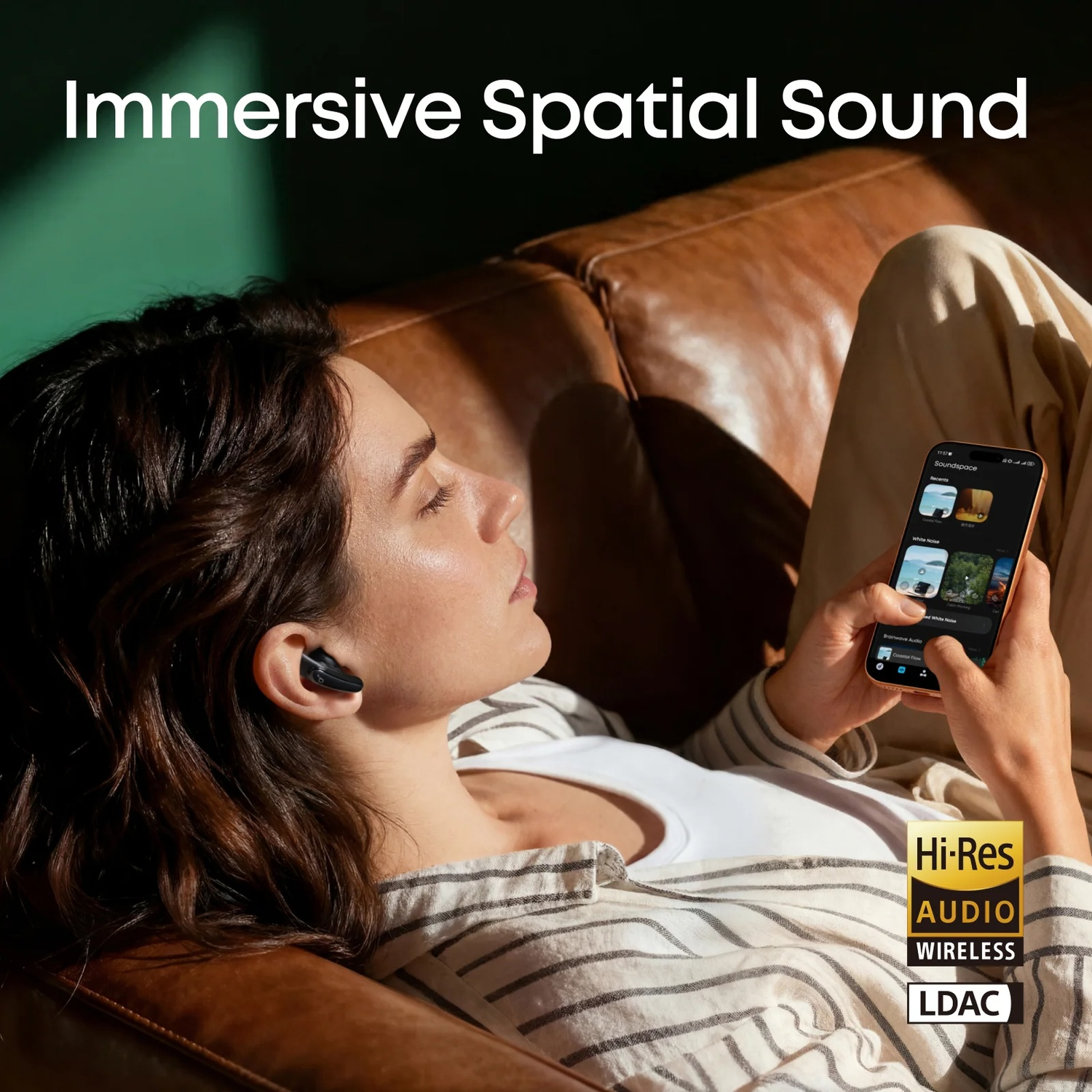 Woman with earbuds relaxes on a brown leather couch, holding a smartphone.