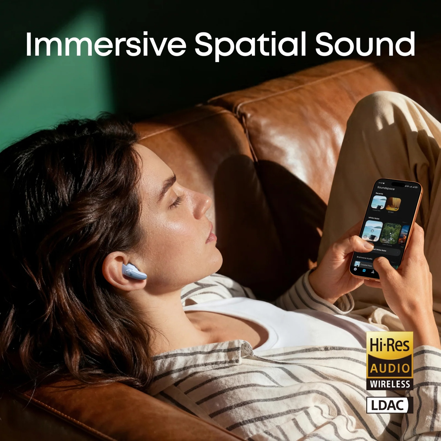 A woman wearing earbuds rests on a brown couch, holding a smartphone.
