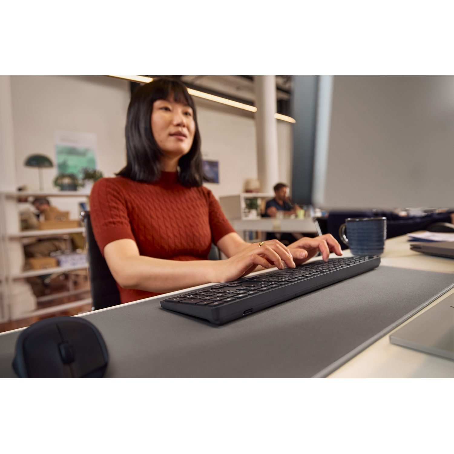 A woman types on a keyboard, with a mouse nearby. An office is in the background.