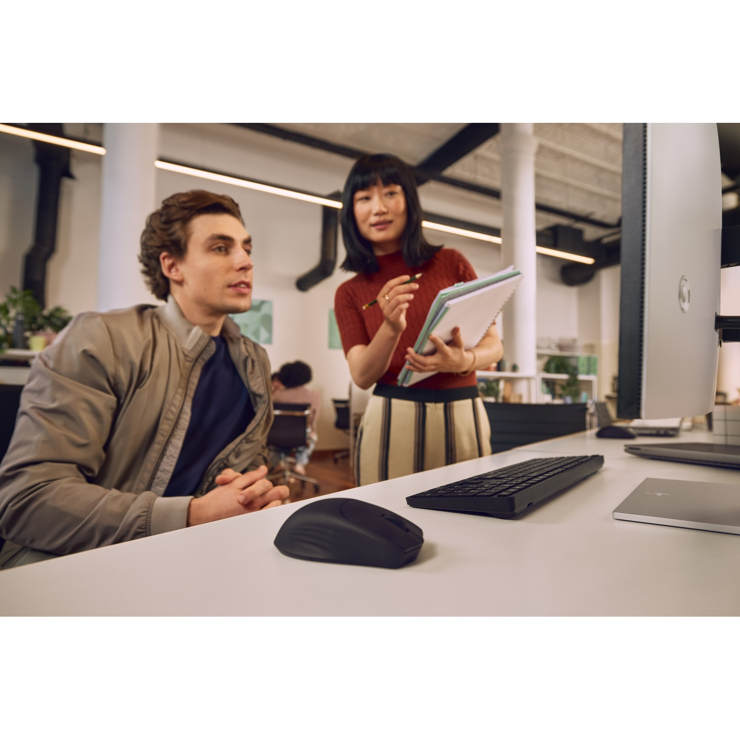 Man and woman at desks with computers and notebooks. Bright office setting.