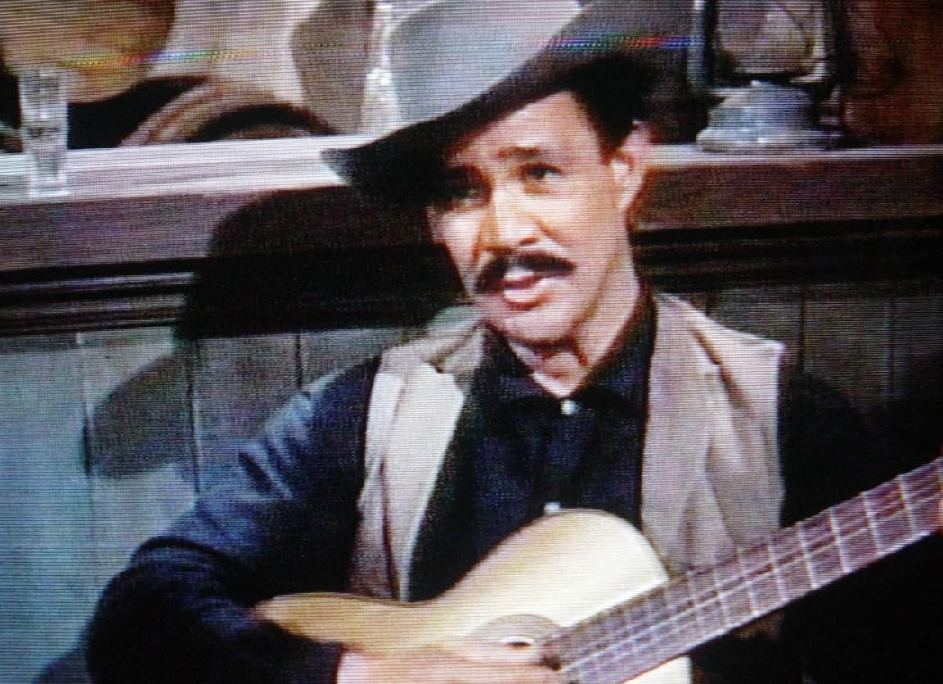 Mann mit Schnurrbart spielt Gitarre in einer Bar. Er trägt Hut, Weste und dunkles Hemd.