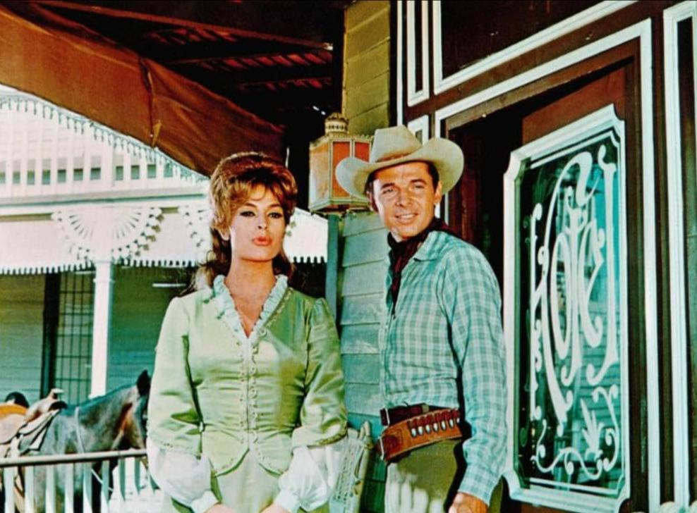 Ein Mann in Cowboykleidung und eine Frau im grünen Kleid stehen vor einem Gebäude im Wilden Westen.