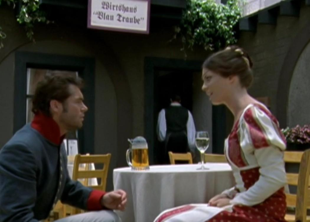Mann in Uniform und Frau in Tracht sitzen am Tisch; im Hintergrund Kellner.