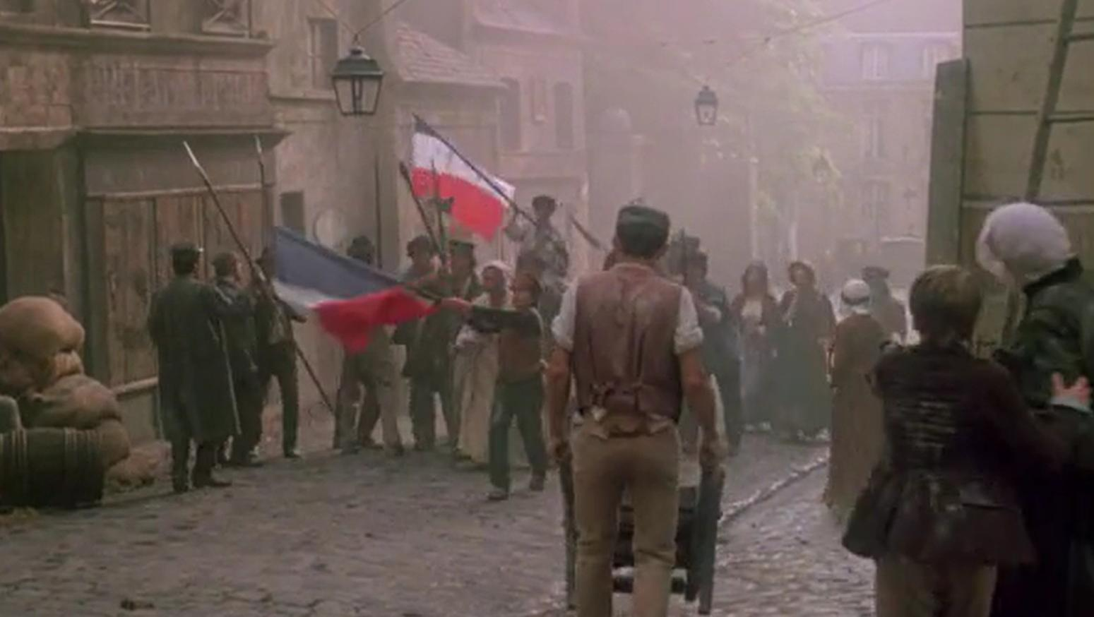 Eine Menge von Menschen in historischer Kleidung schwenkt französische Flaggen auf einer staubigen Straße.