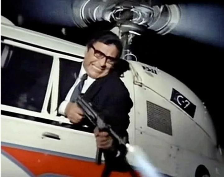 Ein Mann mit Brille hält eine Waffe aus einem Helikopterfenster.