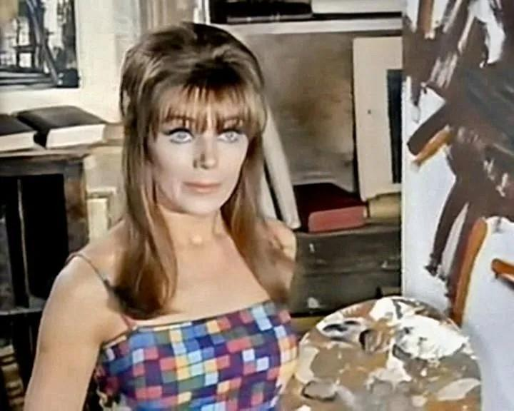 Eine Frau mit gemustertem Kleid und einem Malkasten steht vor einer Leinwand.