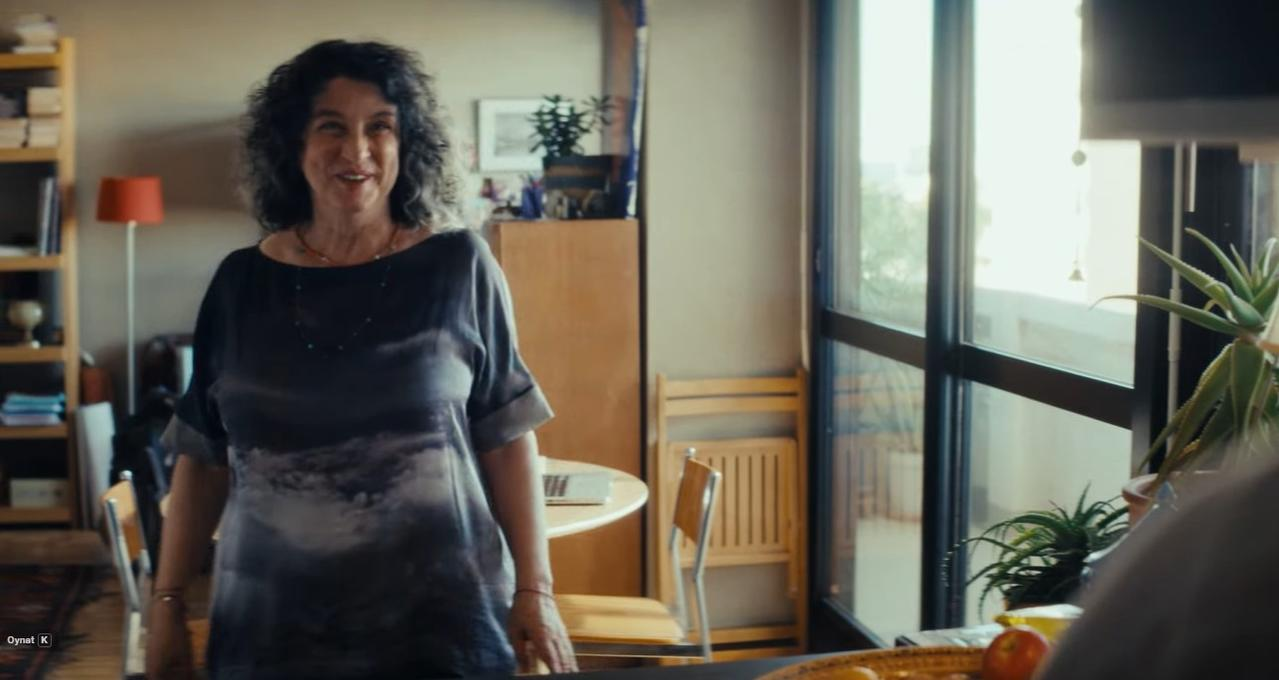Eine Frau mit dunklen Locken steht in einem Raum mit Regalen und Fenstern.