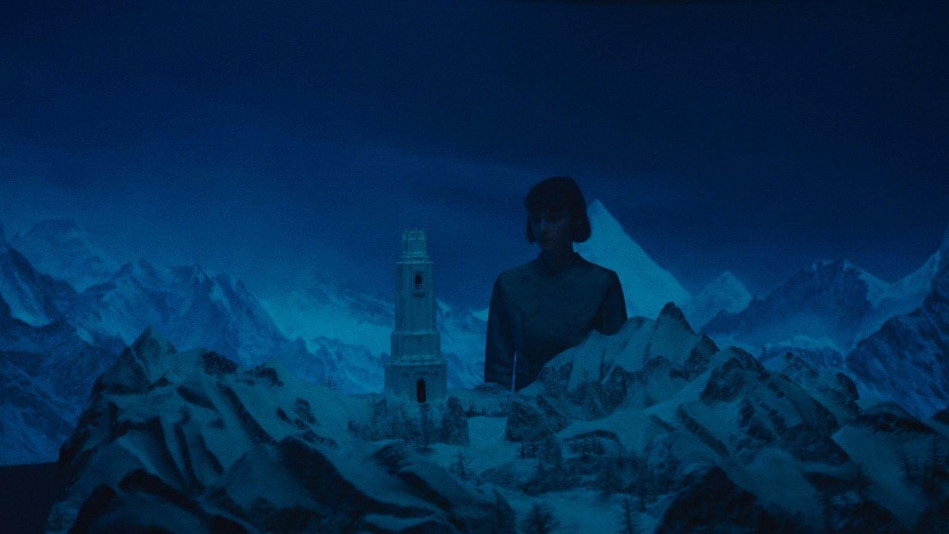 Eine Frau steht vor einem Leuchtturm in einer verschneiten Berglandschaft.
