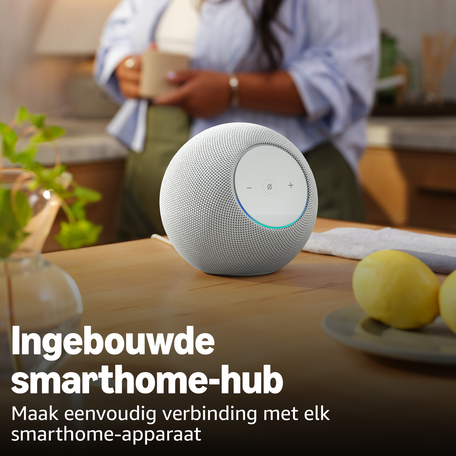 A round, grey smart speaker sits on a wooden table. A person is in the background.