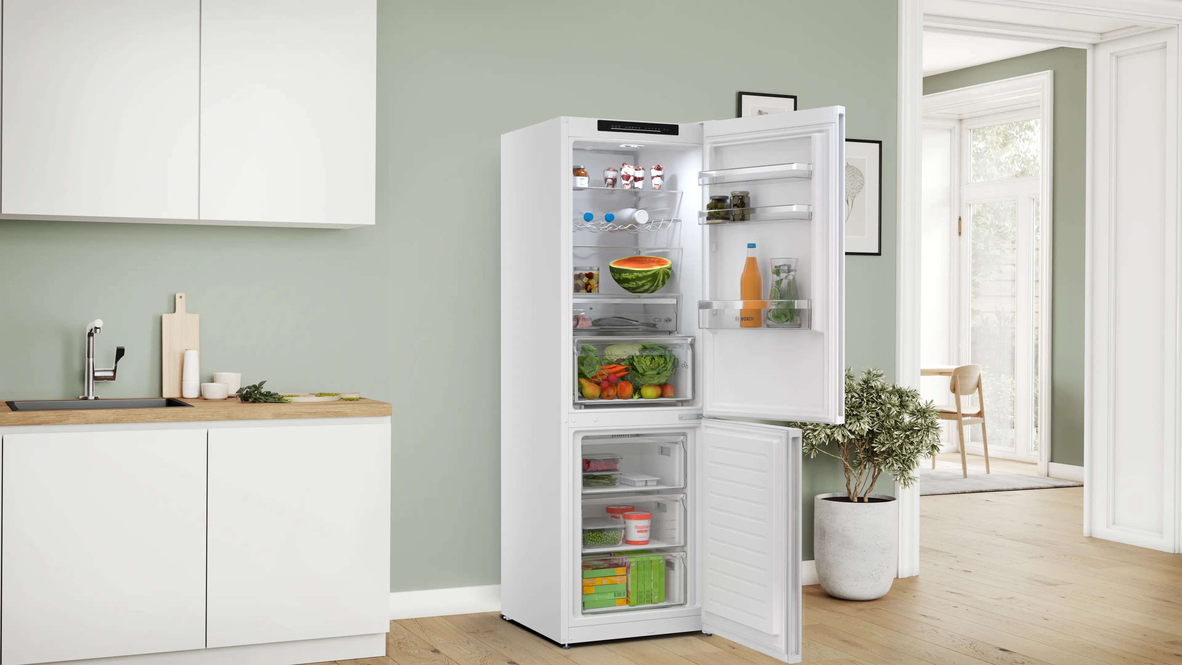 Refrigerador blanco abierto con comida, al lado de una encimera y una planta en maceta.