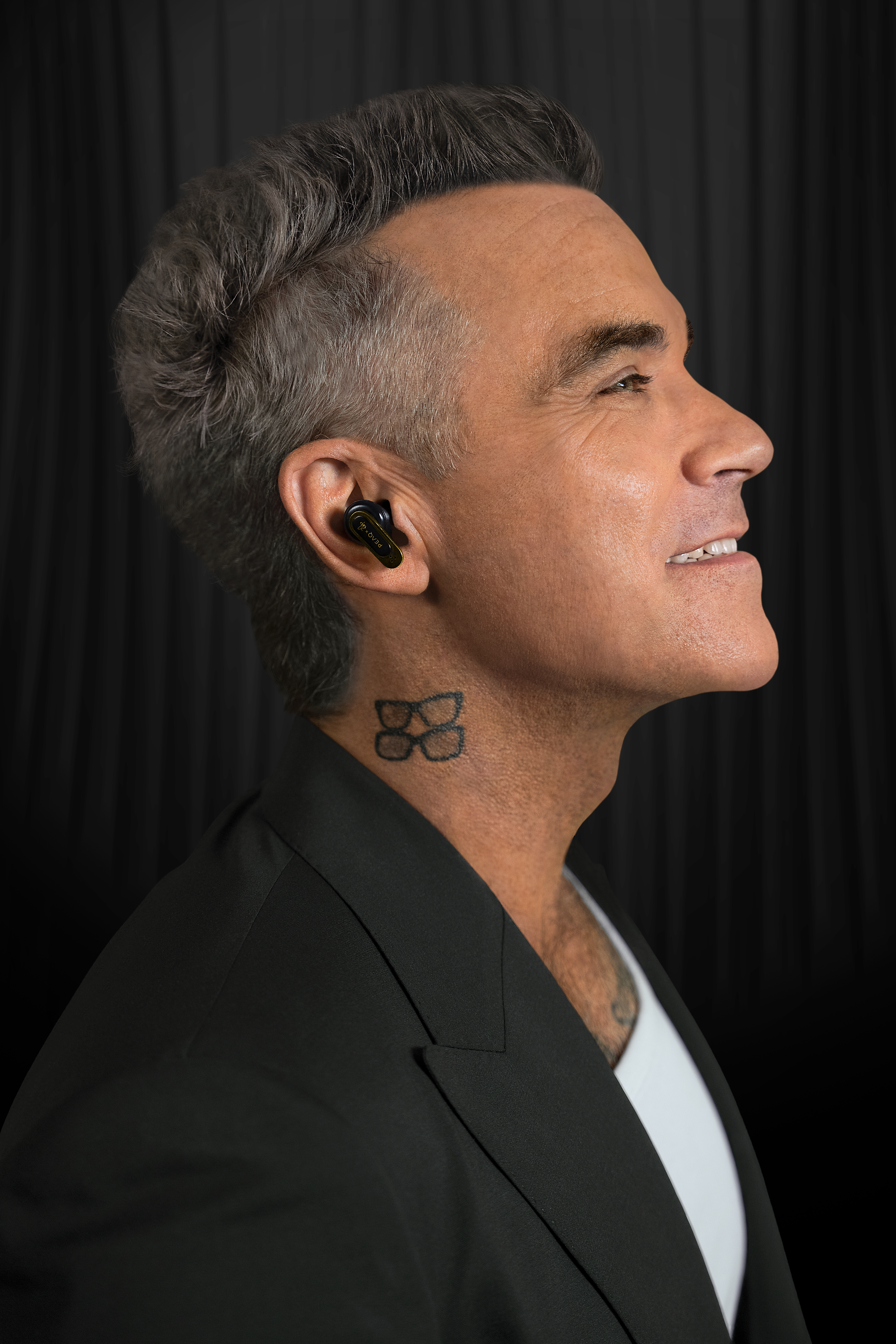 Mann mit schwarzen Ohrhörern und Brillen-Tattoo am Hals, schwarzer Anzug.