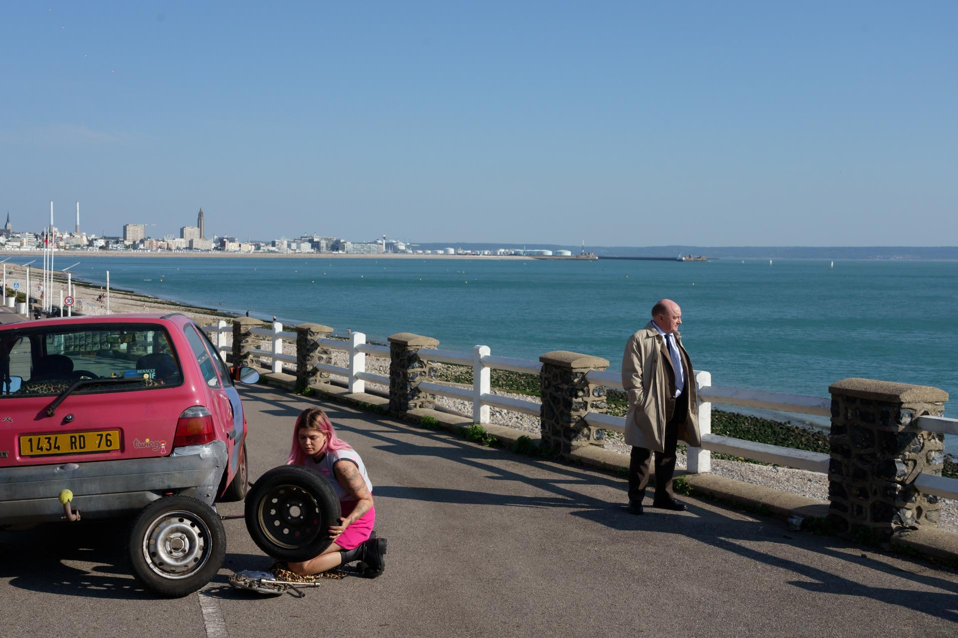 Eine Frau mit pinken Haaren wechselt das Rad an einem roten Auto. Ein Mann steht daneben.