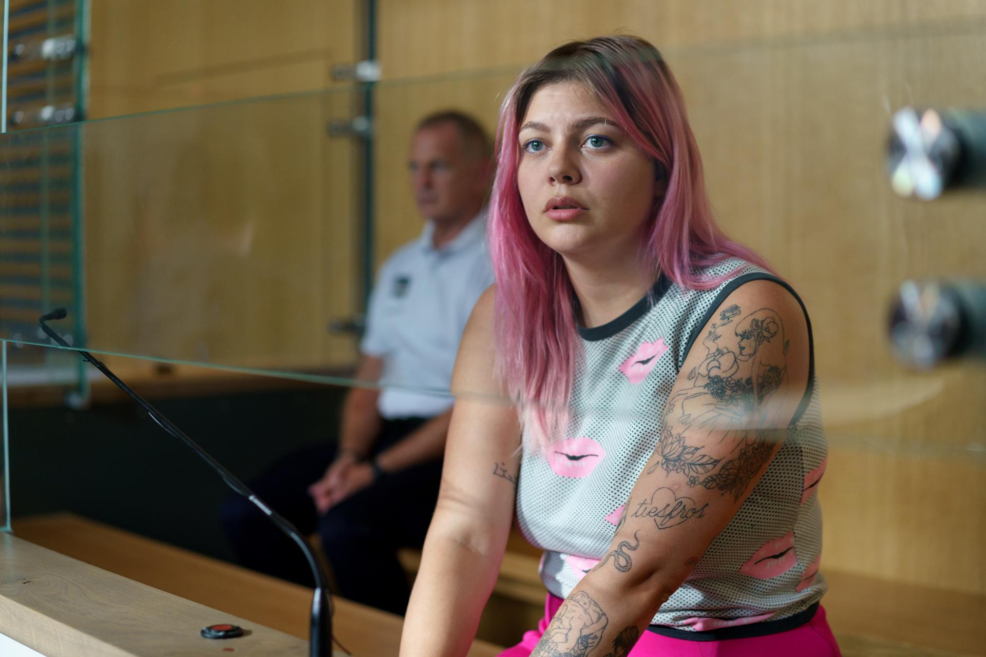 Eine Frau mit pinken Haaren und Tattoos sitzt hinter Glas. Ein Mann im Hintergrund.