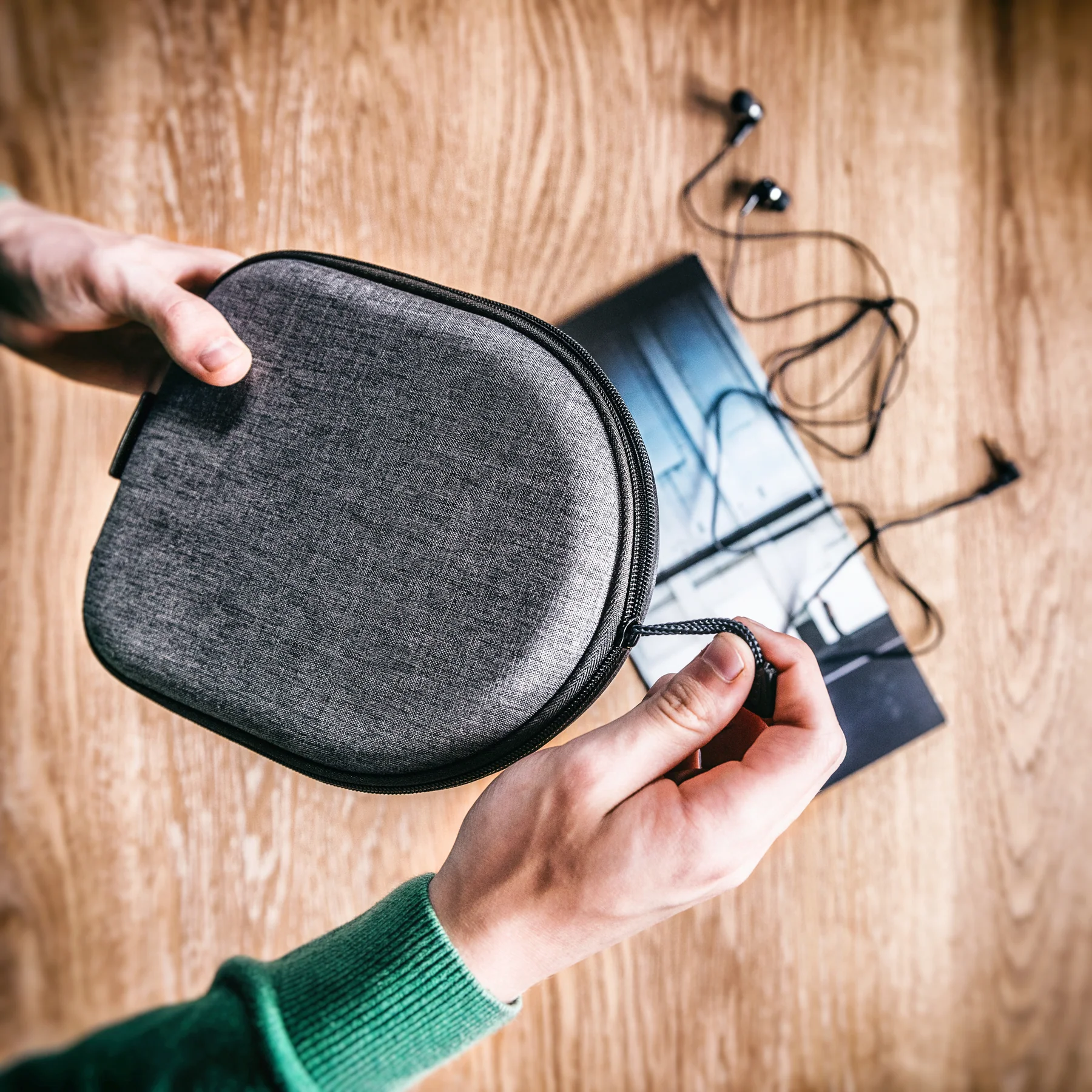 Mani aprono una custodia grigia con zip su tavolo in legno. Auricolari accanto.
