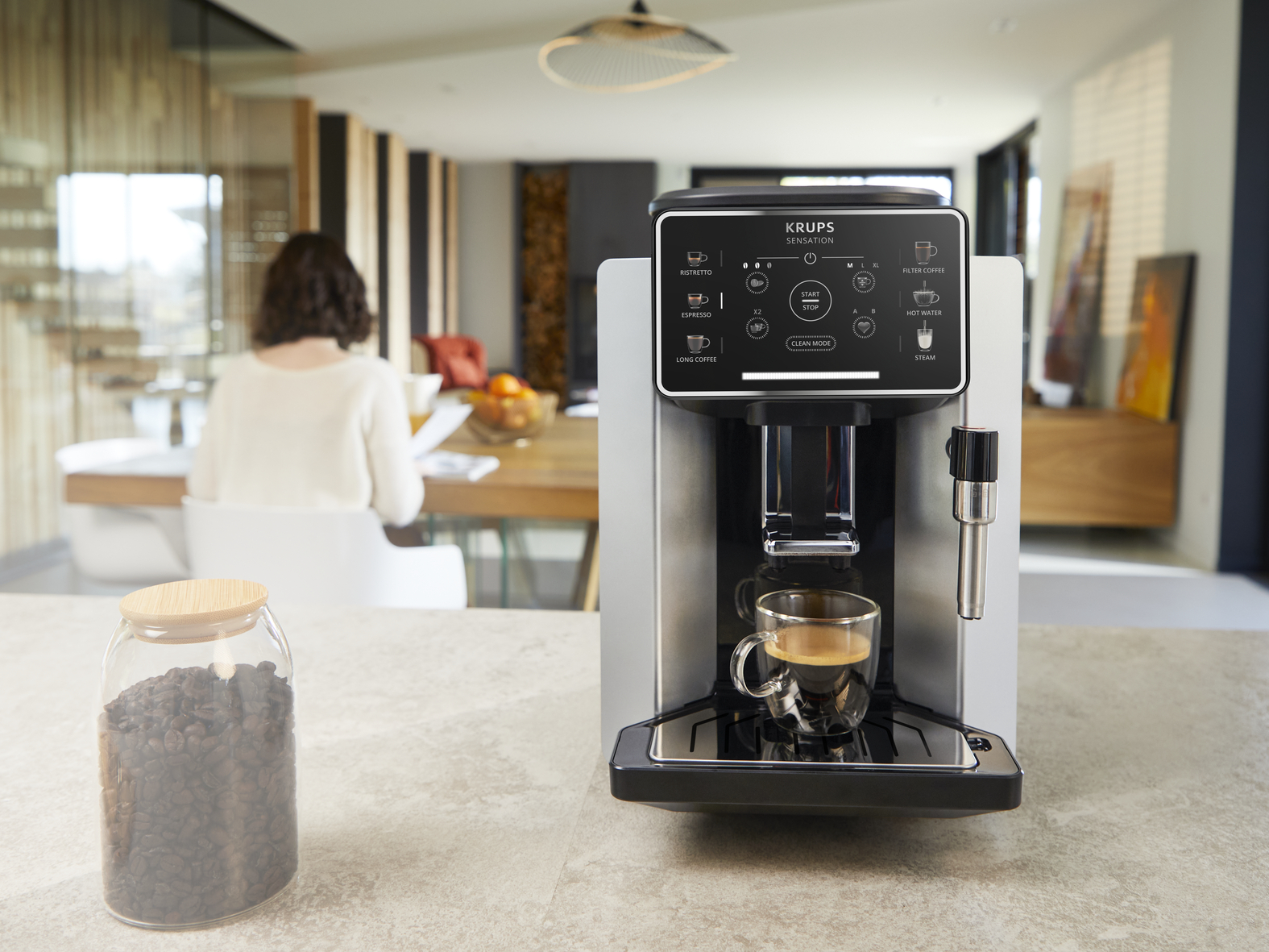 Krups automatic coffee machine brewing espresso in a glass cup. Coffee bean jar in foreground.