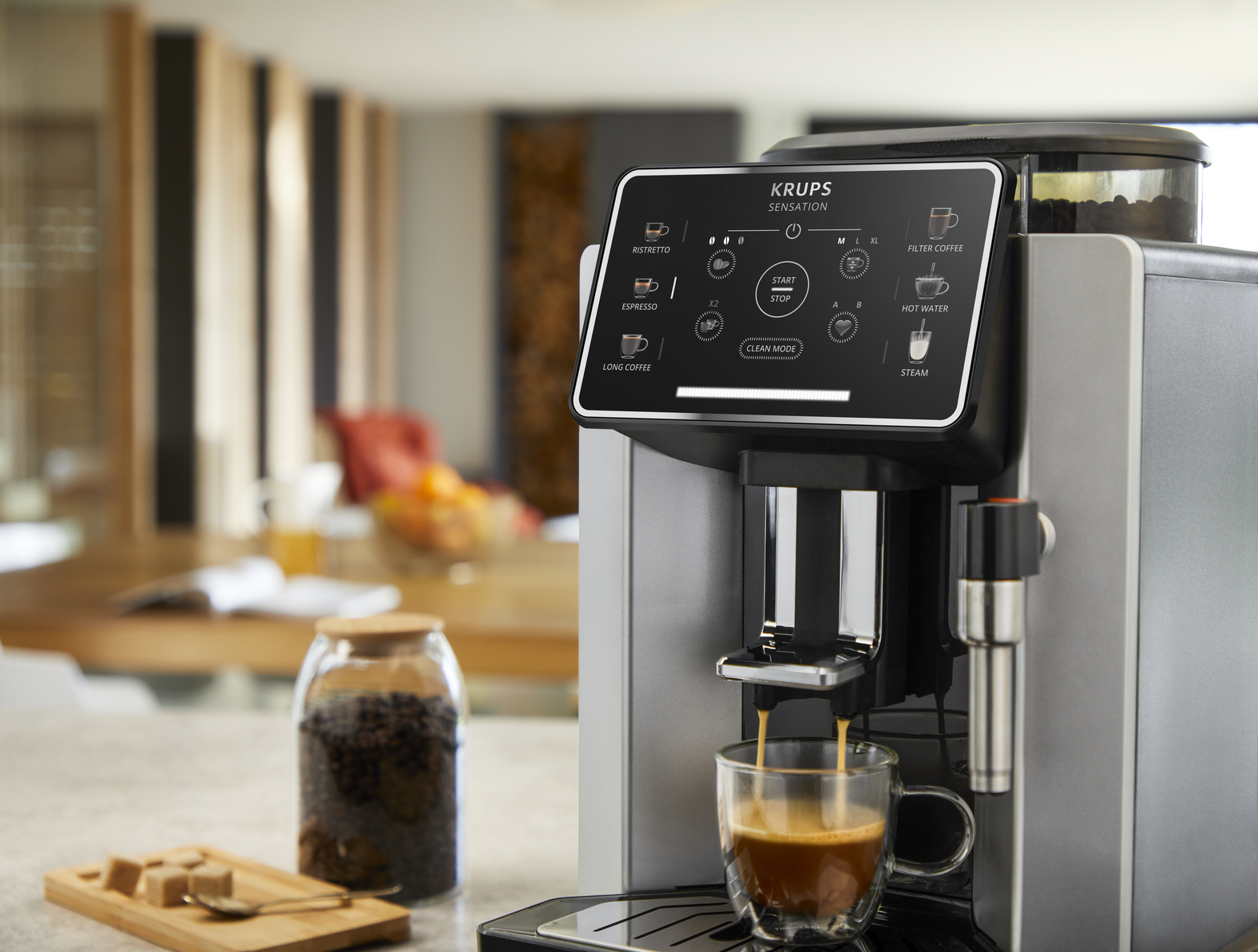 Krups coffee machine brews espresso into a cup. Coffee beans and sugar cubes nearby.