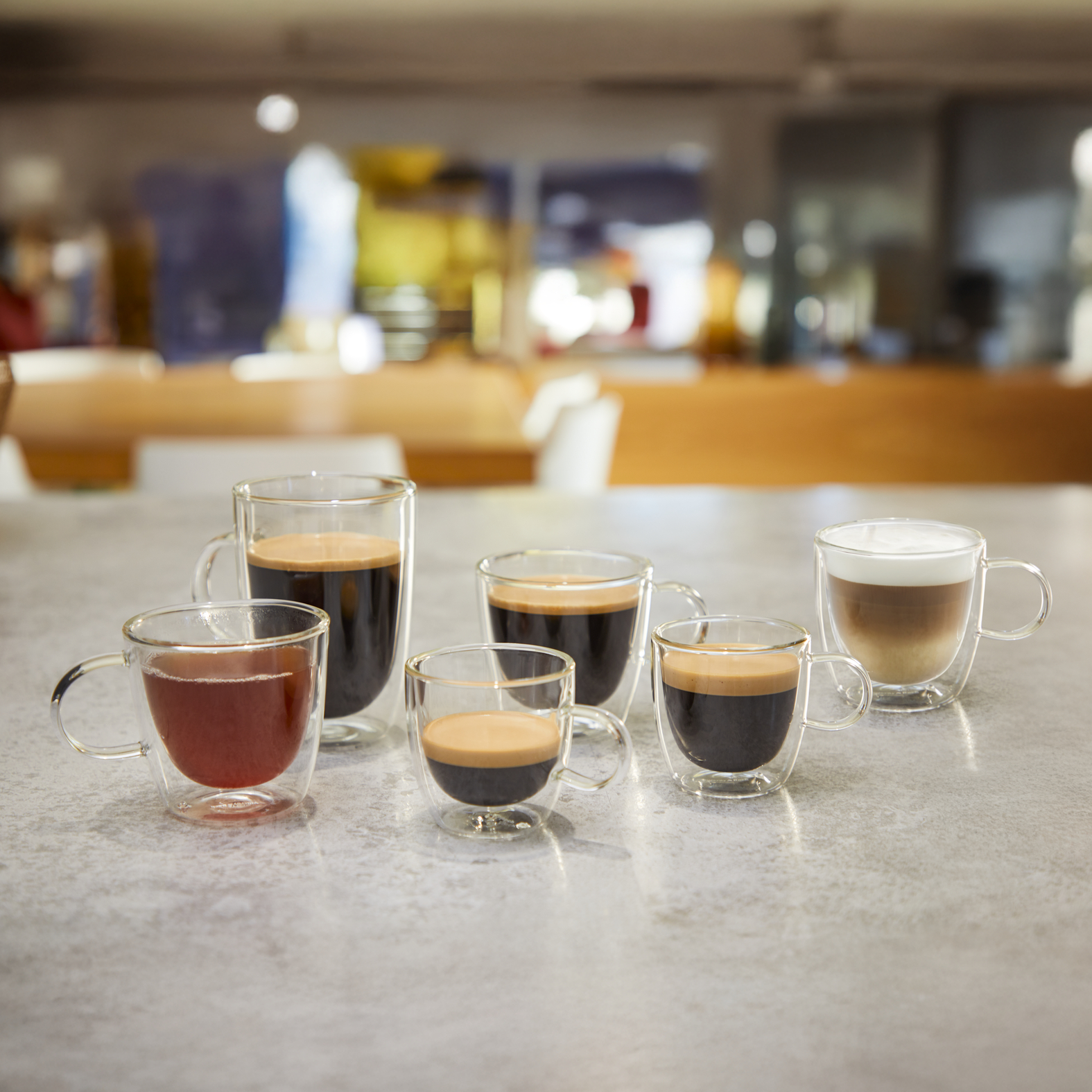 Six glasses with coffee drinks and one red drink on a table.