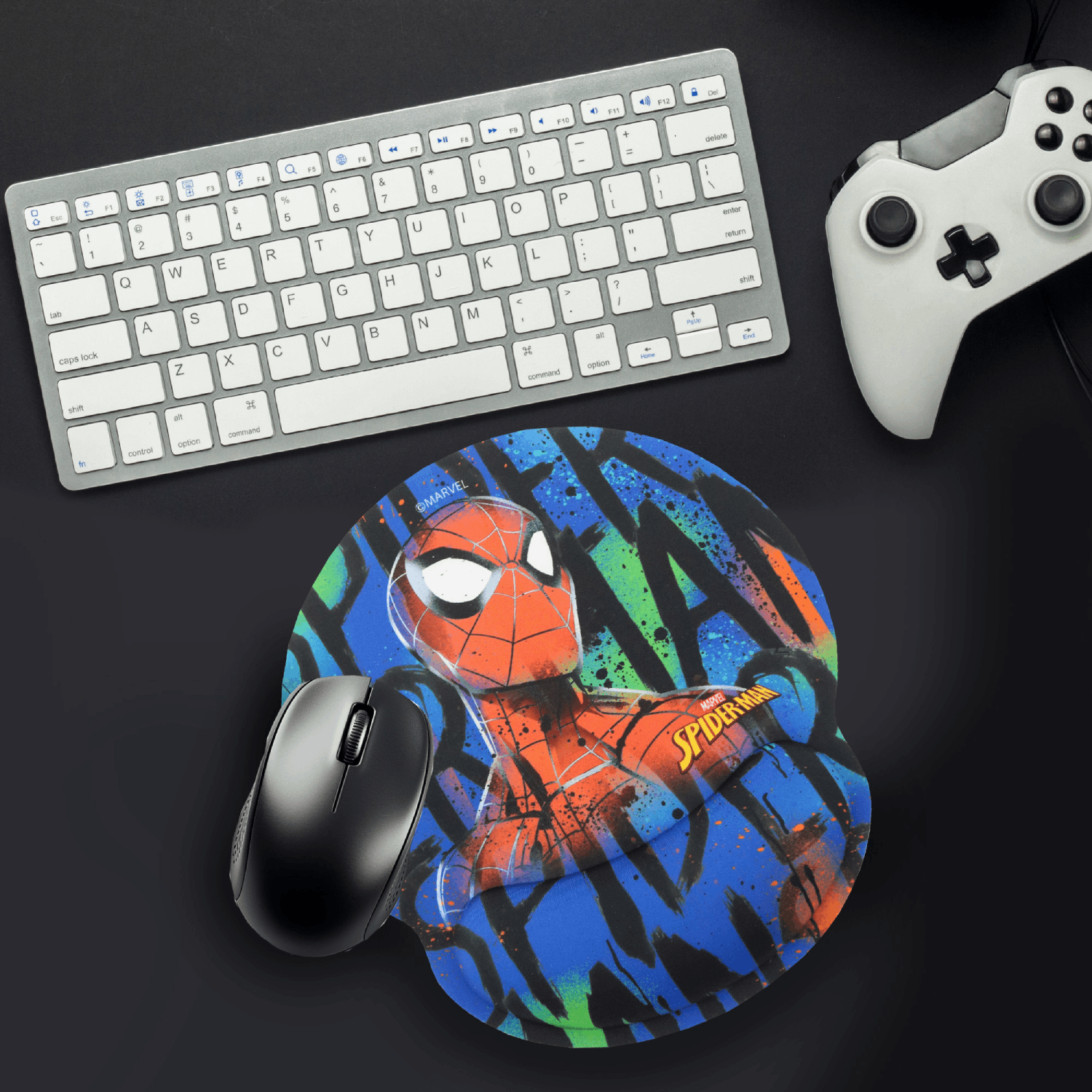 Un ratón gaming negro descansa sobre un reposamuñecas de Spiderman, con teclado y mando cerca.