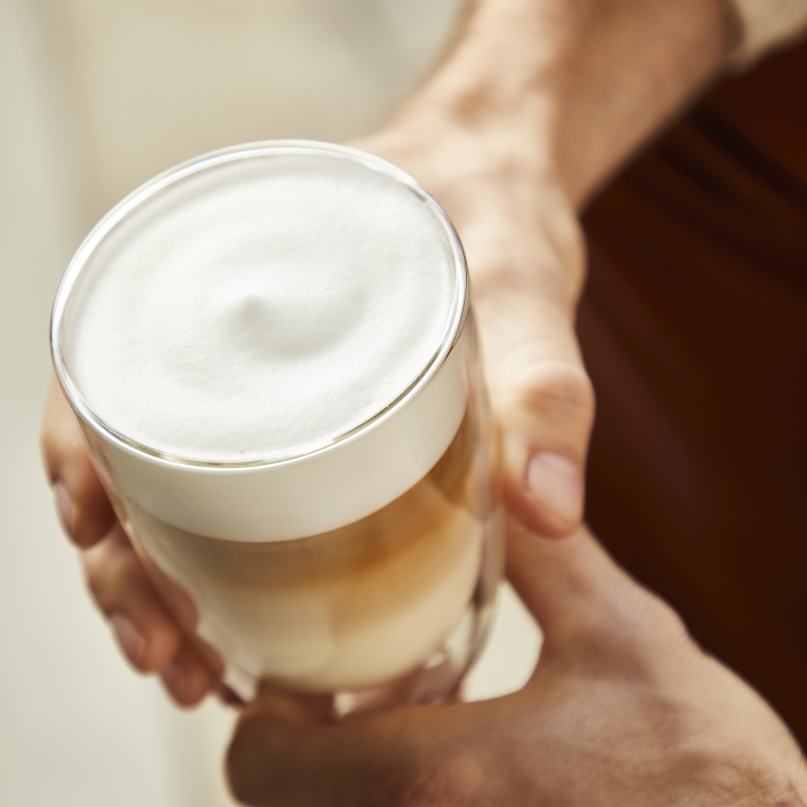 Handen houden een glas met gelaagde koffie en melkschuim vast.