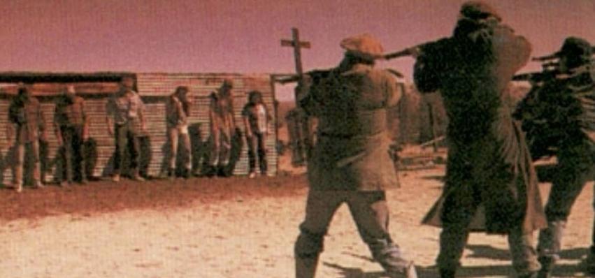 Männer mit Gewehren zielen auf eine Gruppe von Menschen vor einer Holzmauer.