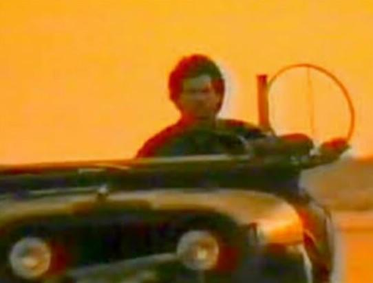 Mann in einem Jeep mit einem Rad im Vordergrund, orangefarbener Himmel.