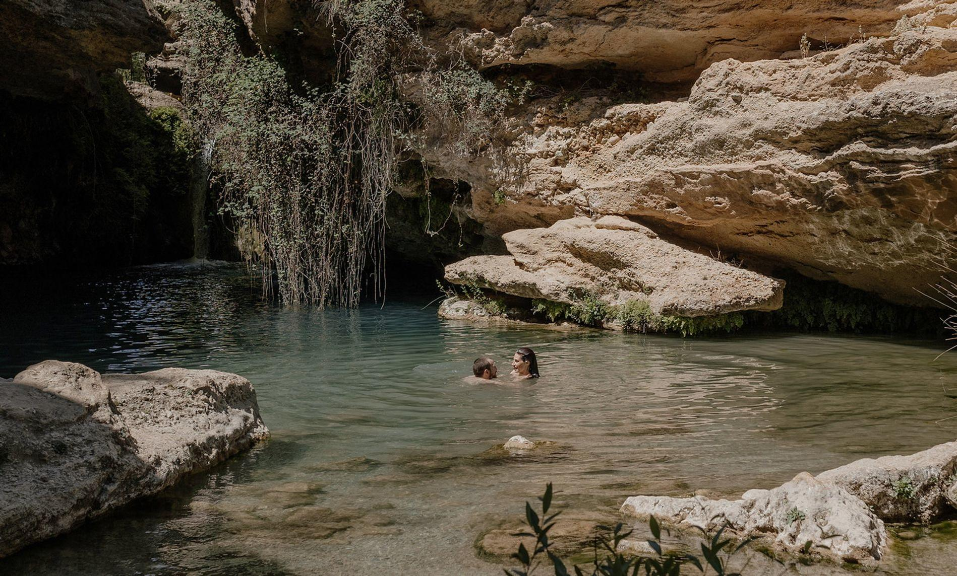 Zwei Personen schwimmen in einem klaren See mit Felsen und Vegetation.