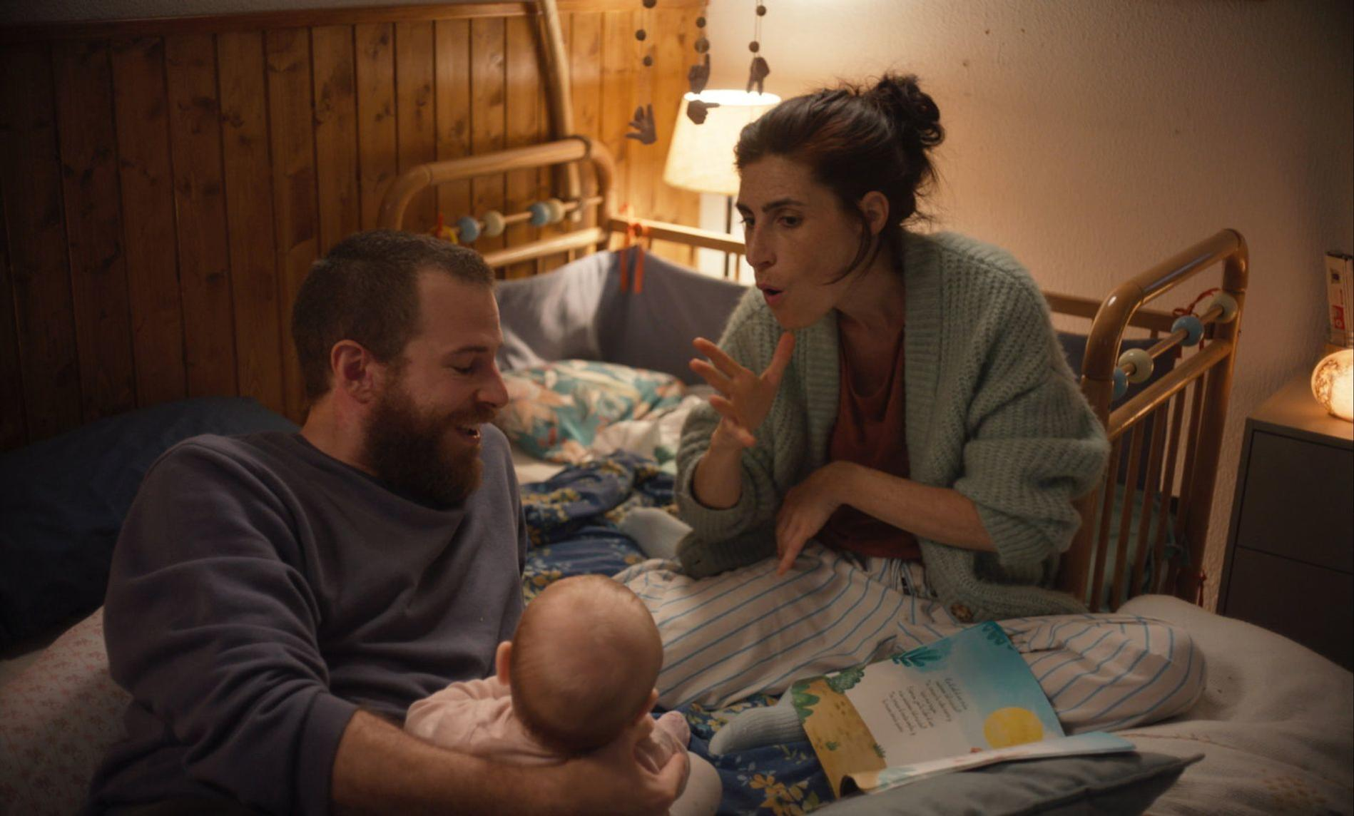 Eltern lesen Baby im Bett eine Geschichte vor, ein Kinderbett im Hintergrund.