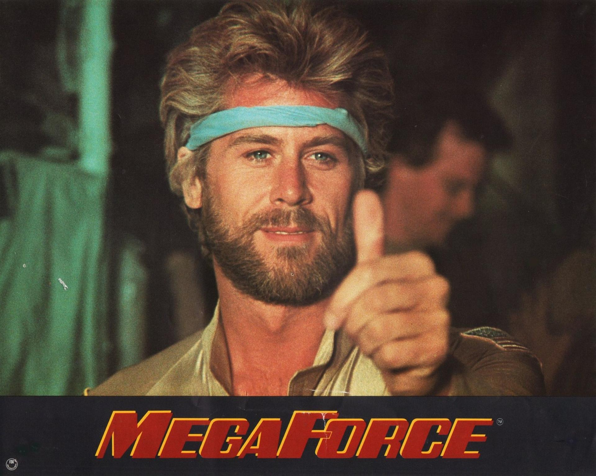 Ein Mann mit Bart und blauem Stirnband gibt Daumen hoch. Der Titel "MEGA FORCE" ist unten.
