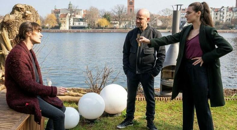 Drei Personen stehen am Wasser, Gebäude im Hintergrund.
