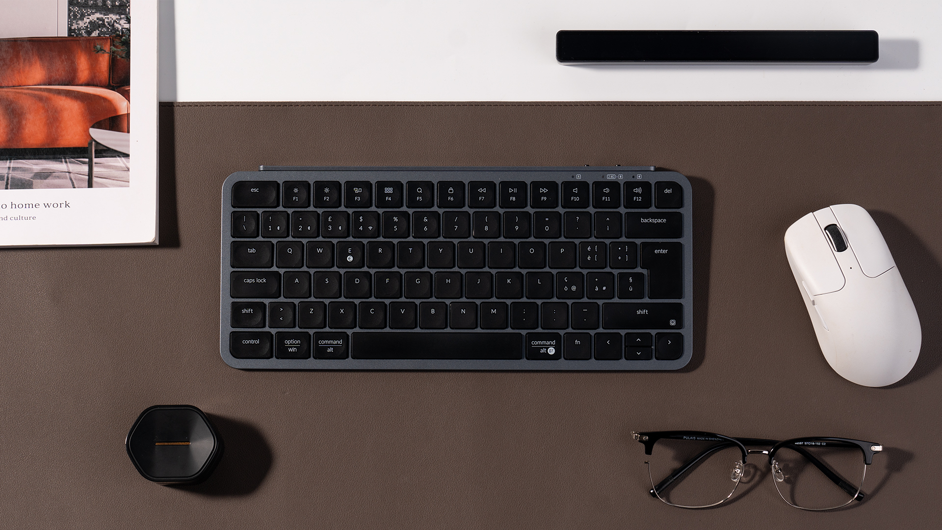 Ein graues Keyboard, weiße Maus, Brille und ein Buch auf braunem Untergrund.