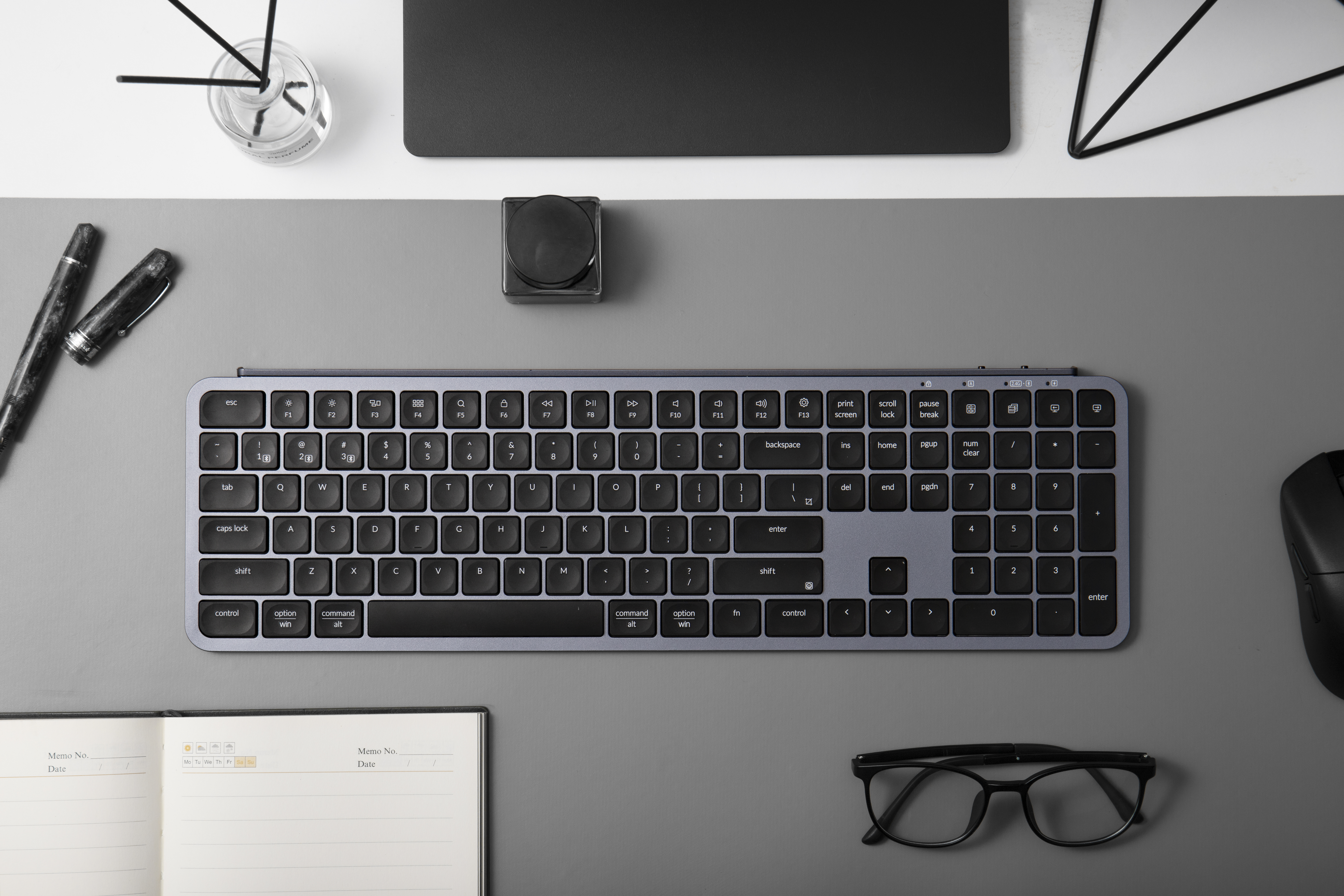 Ein dunkles Tastatur-Setup mit Maus, Brille und Notizbuch auf grauem Hintergrund.