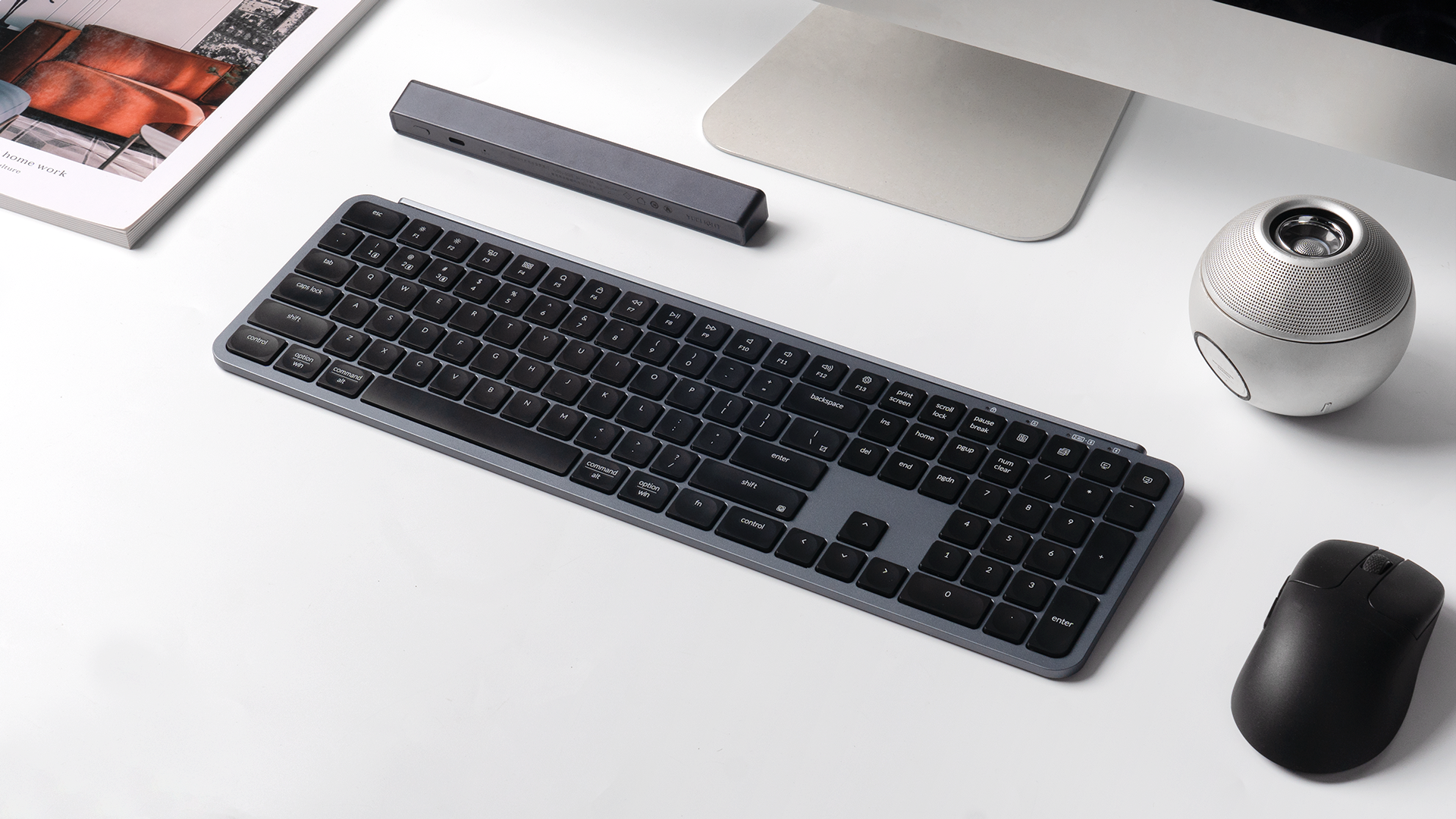 Graue Tastatur, schwarze Maus, runder Lautsprecher und Buch auf weißem Tisch.