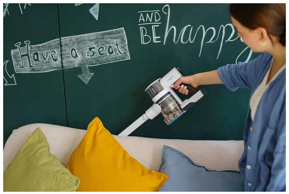 Eine Frau saugt mit einem Handstaubsauger ein Sofa ab. An der Wand steht „Have a seat“.