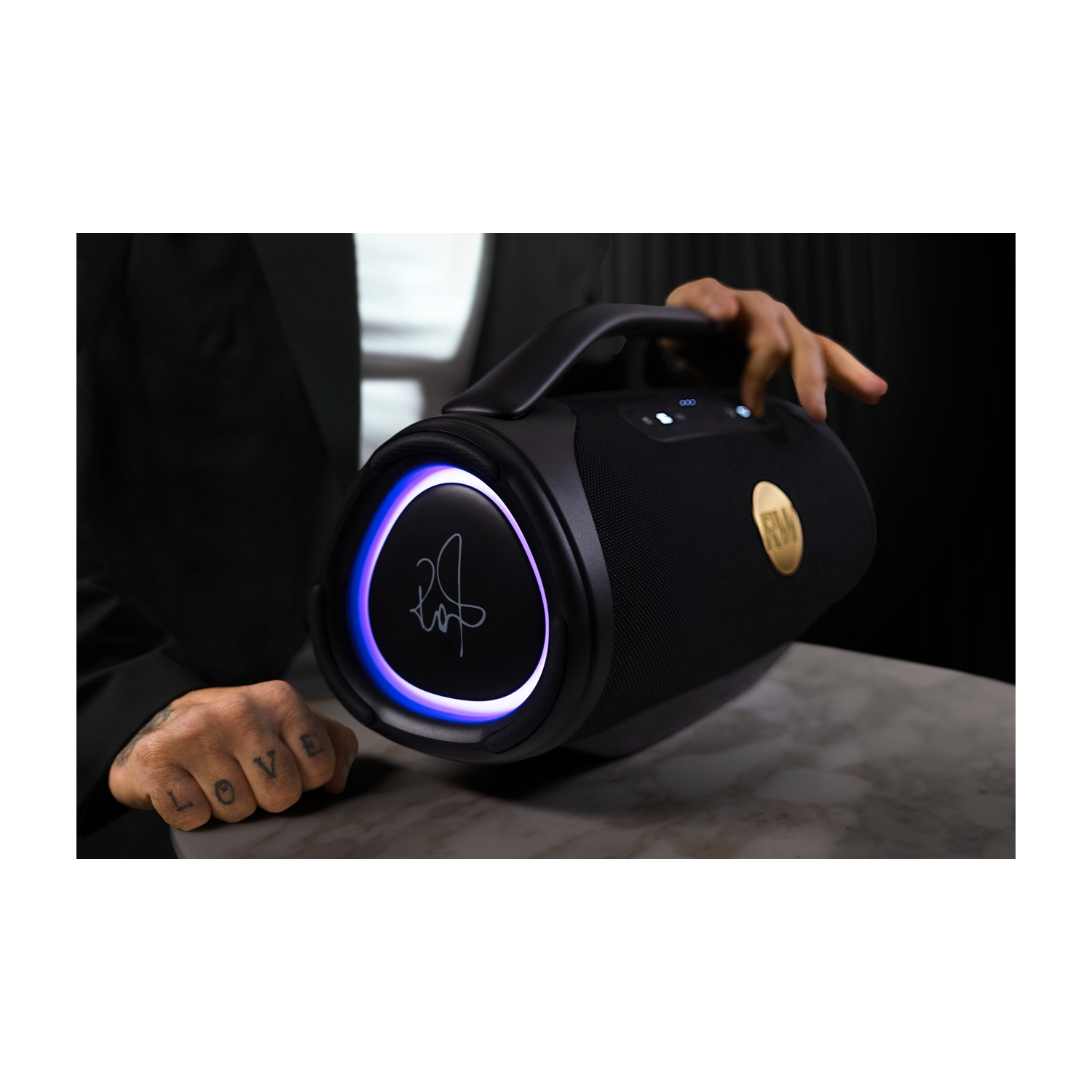Un altoparlante nero con anello luminoso blu, tenuto da una mano tatuata.