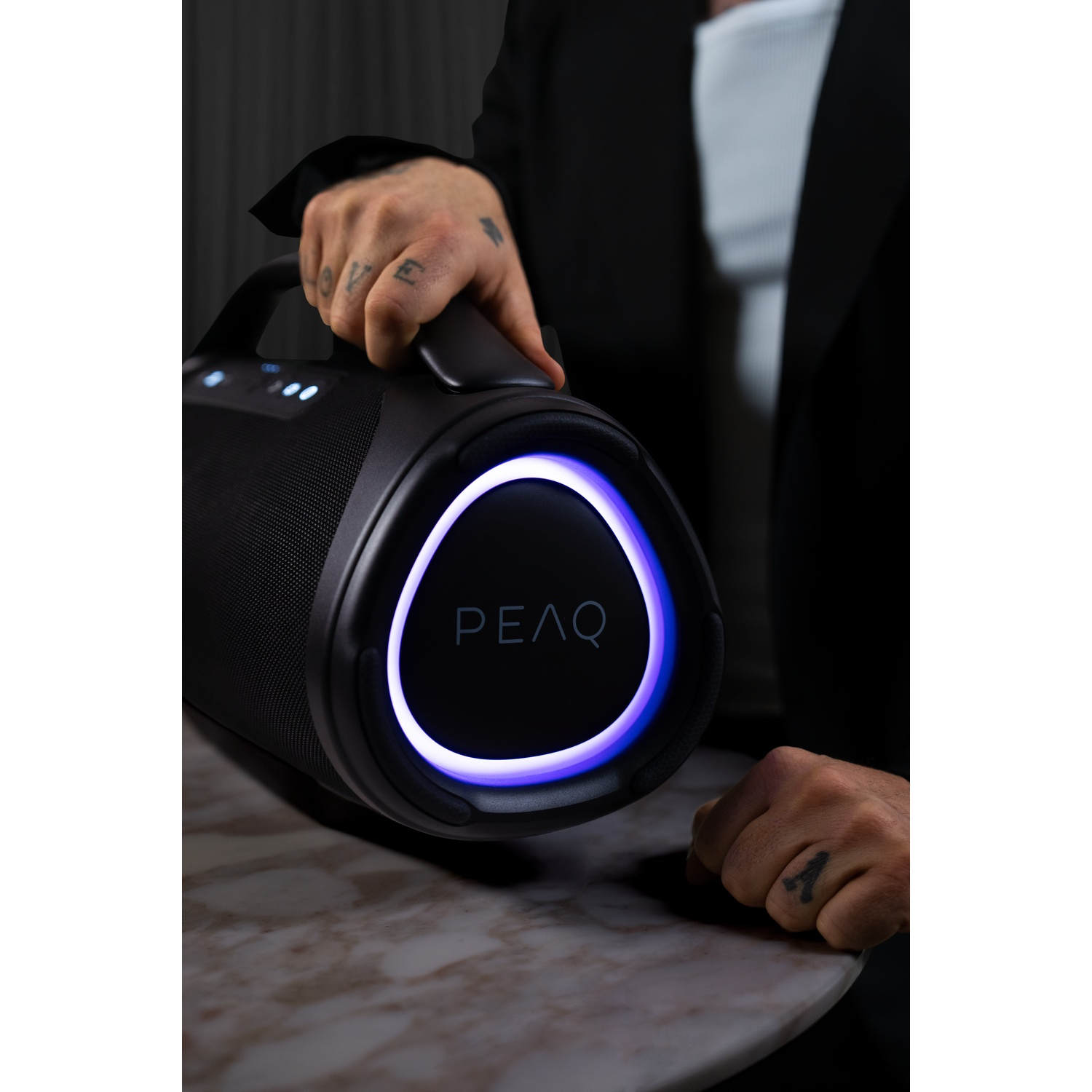 Una persona tiene un altoparlante nero con anello luminoso blu. Il logo PEAQ è visibile.