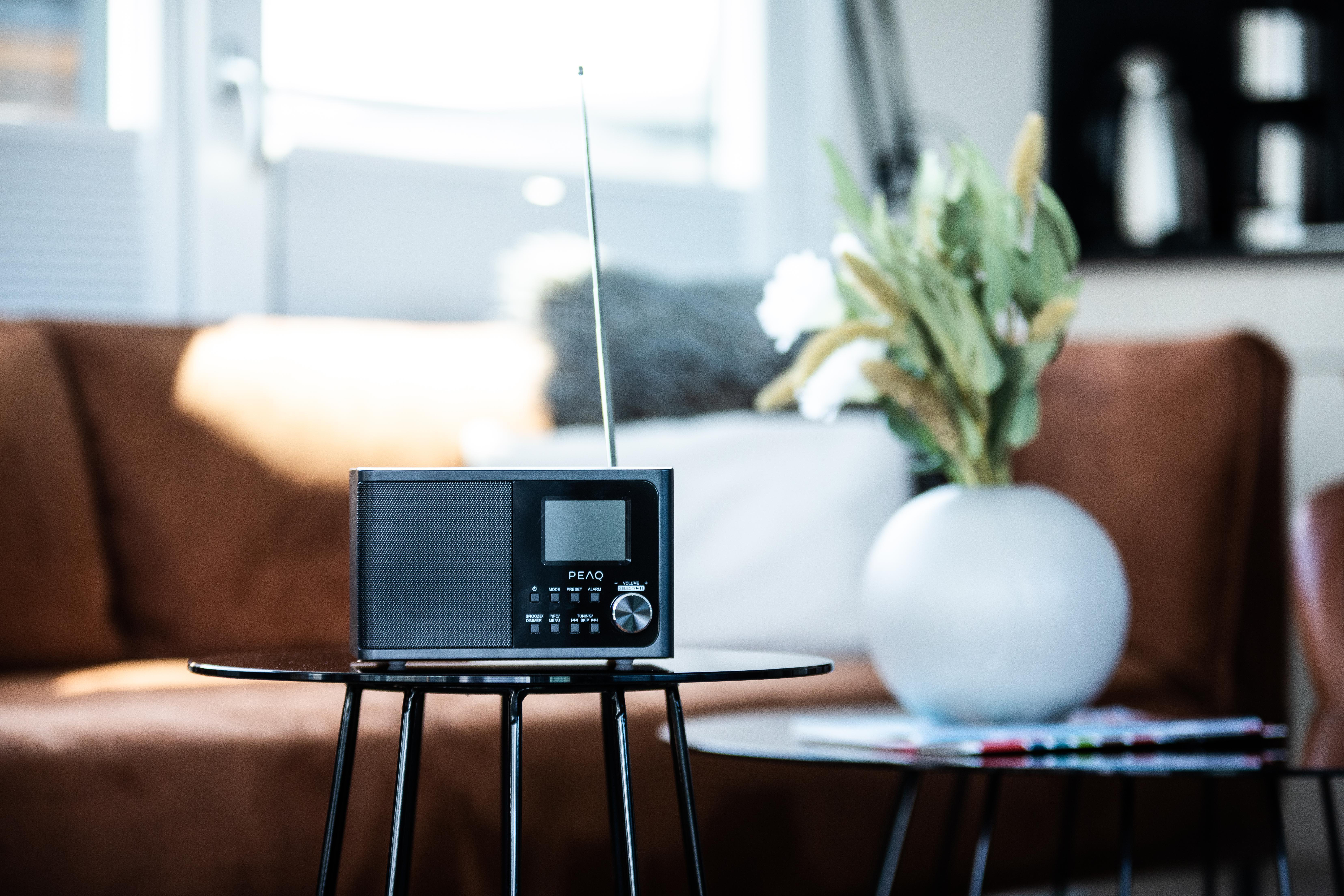 Ein schwarzes Radio auf einem Glastisch vor einem braunen Sofa, daneben eine weiße Vase.